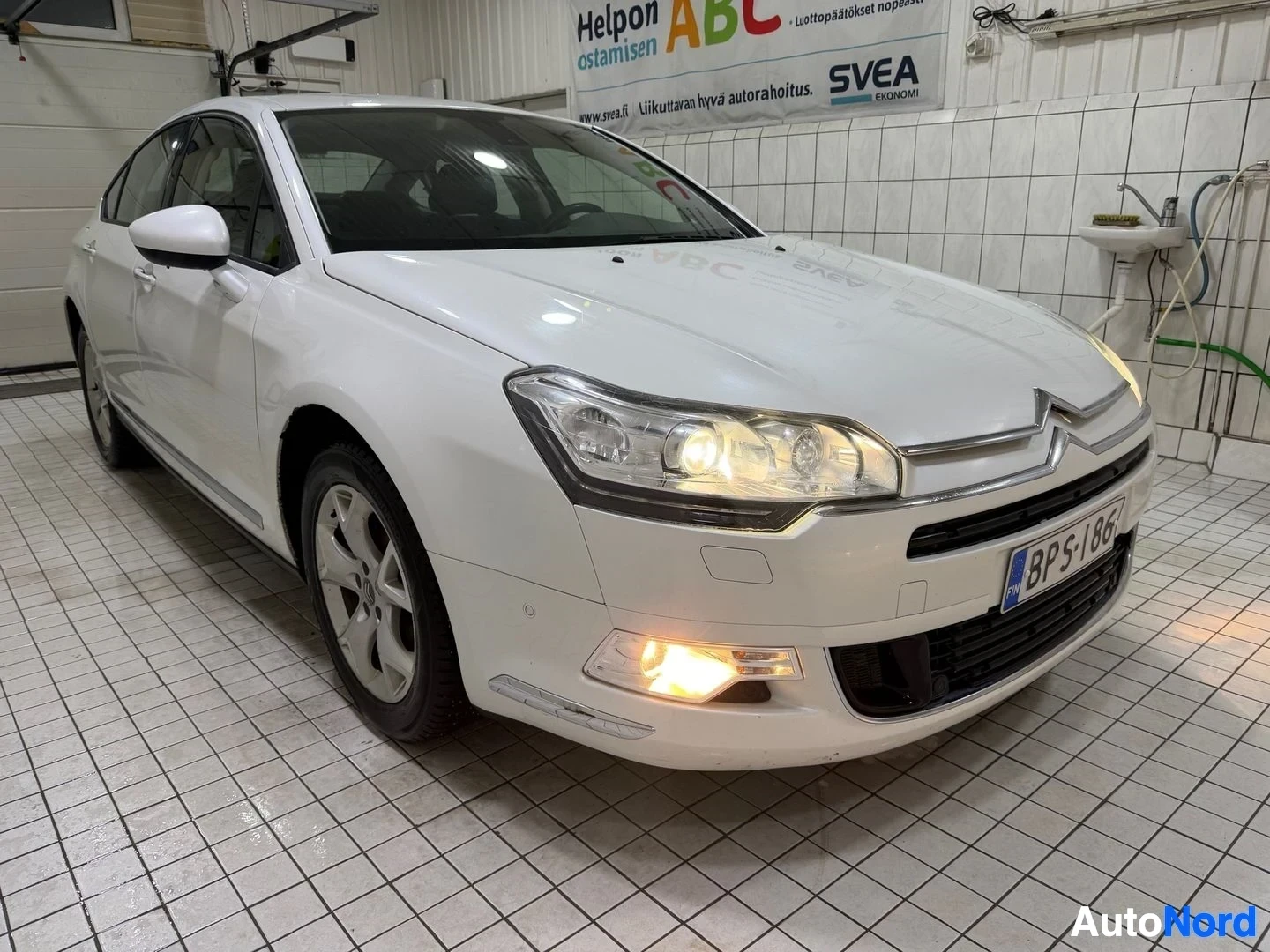 Etuviisto, nokka vasemmalle: Valkoinen Citroen-sedan kylmävalaistulla hallilla, oikealle käännettynä, edusta ja oikea sivu näkyvissä.