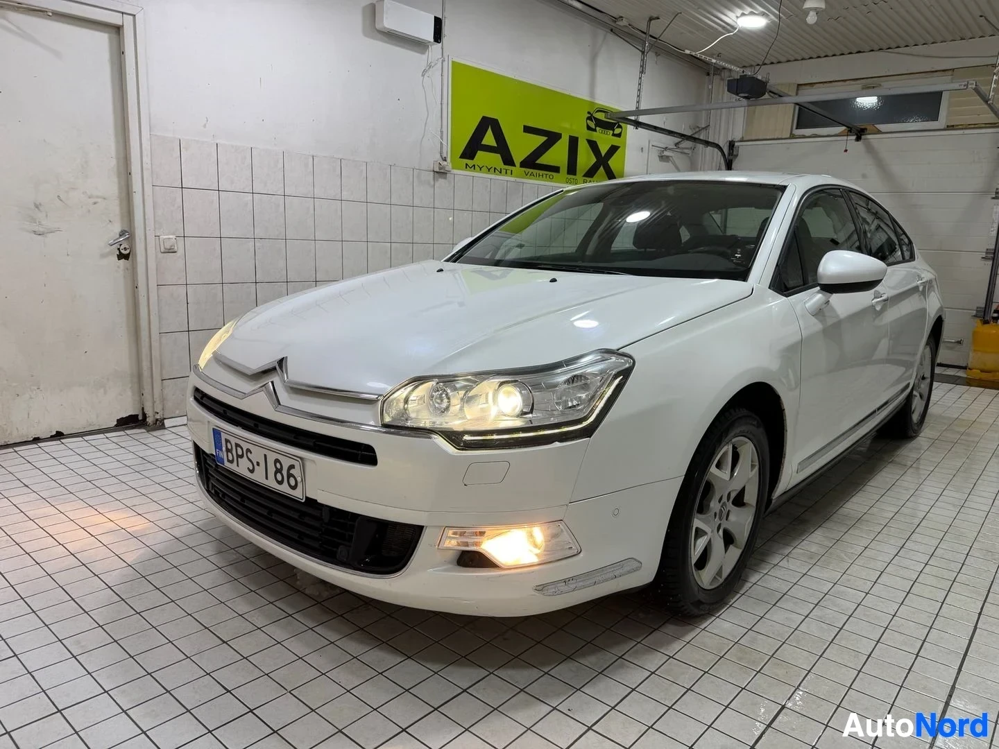 Etuviisto, nokka oikealle: Valkoinen Citroen-sedan vasemmalla kolmella neljäsosalla, vasemmalla puolella ja etupuolella näkyvissä hallissa.