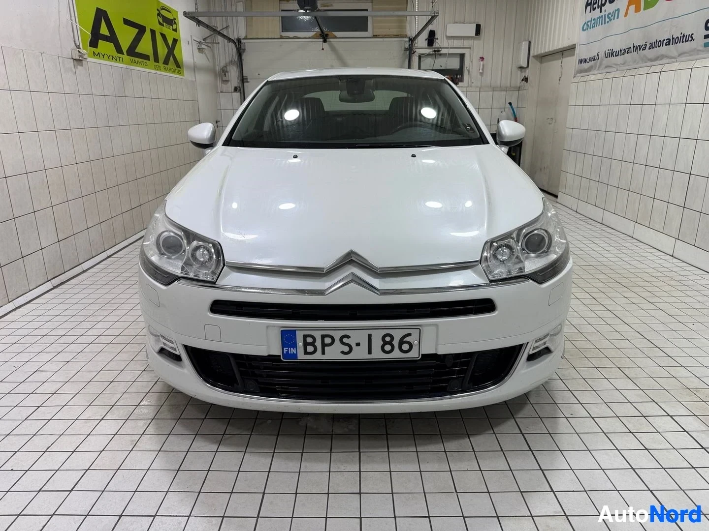 Edestä: Valkoinen Citroën-sedan edestäpäin kuvattuna sisäkatsomohallissa. Rekisterikilvessä lukee BPS186. Auto on suoraan katsojaa kohti.