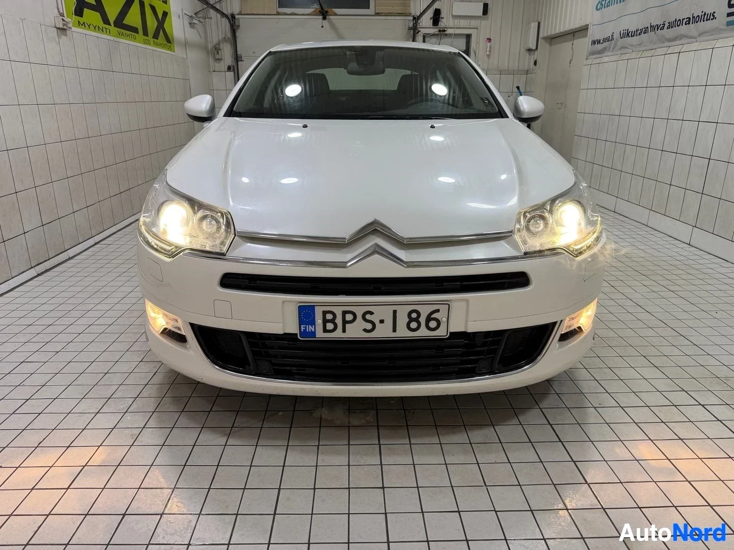 Edestä: Valkoinen Citroen-sedan suoraan edestä katsottuna hallissa, molemmat etuvalaistut palaen.