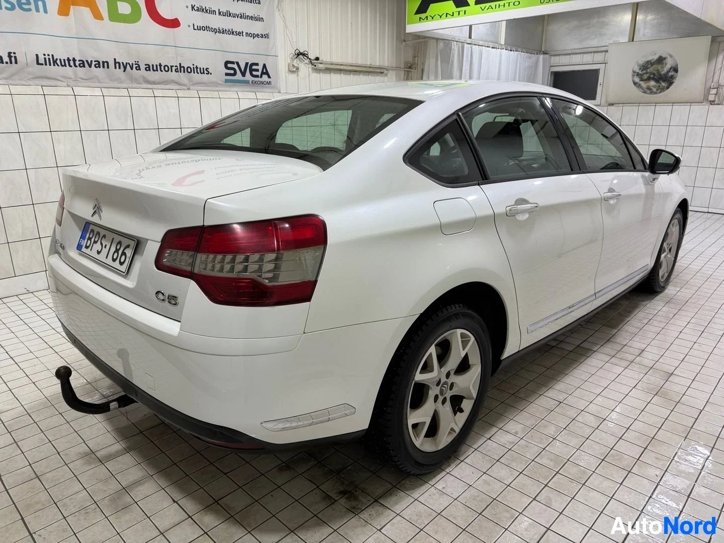Sivulta: Valkoinen Citroen C5 sedan näkyy oikealta sivulta, sisätiloissa. Rekisterikilpi näkyy. Takaovi on auki. Musta vetosilmukka on näkyvissä