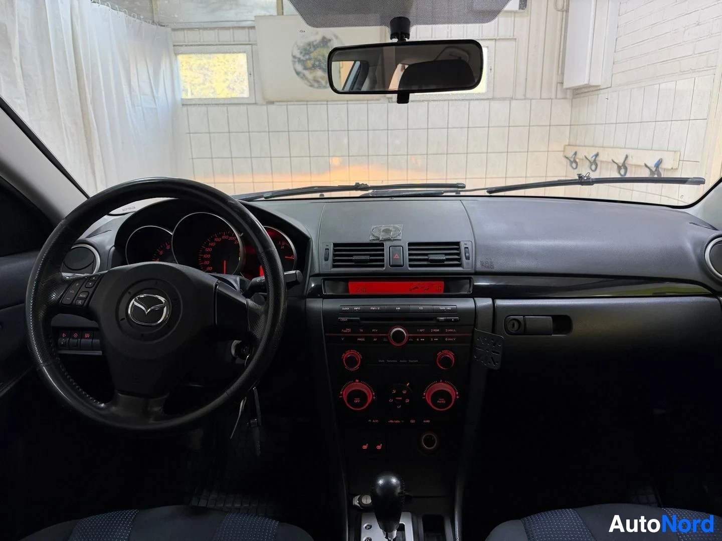 Ohjaamo: Mazda 3 -auton kuljettajan puoleinen näkymä instrumenttipaneelistä, Mazda-merkillä varustettu ohjauspyörä, kaksi pyöreää mittaria punaisilla merkeillä, pääsäänt