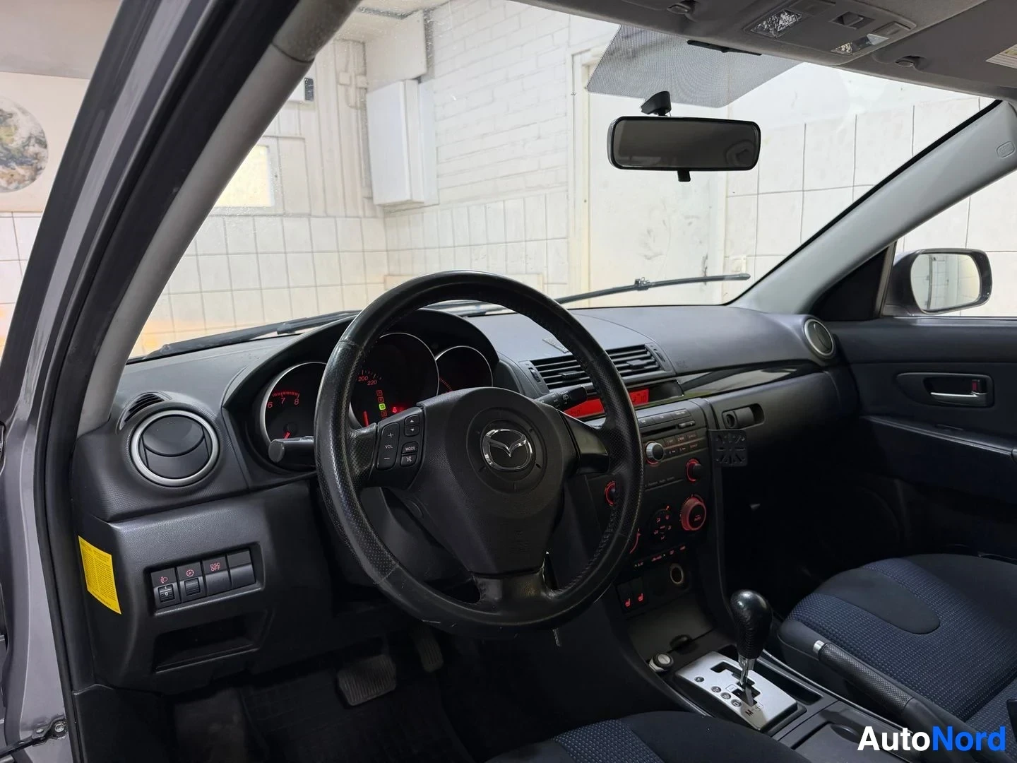 Etuistuimet: Mazda 3 -auton kuljettajan puoleinen etupenkki sisäkuva, Mazda-merkki ohjauspyörässä, musta nahkainen pää-ohjaus, näkyvissä ilmastoinnin säätöyksikkö punaisen v