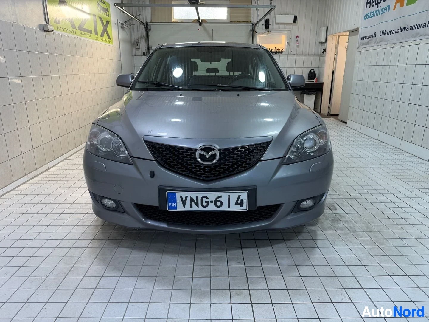Edestä: Mazda 3 -auto edestä katsottuna autotallin sisällä, rekisterikilvessä, Suomen lippu kilvessä, harmaa väri näkyvissä