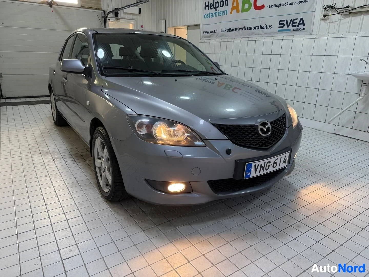 Edestä: Auto etupuoli edestä: hopea väri, Mazda-merkki grillissä, valkoisen väriset vanteet, rekisterikilvessä, pois päältä kääntyneet ajovalot, hallissa sisäll
