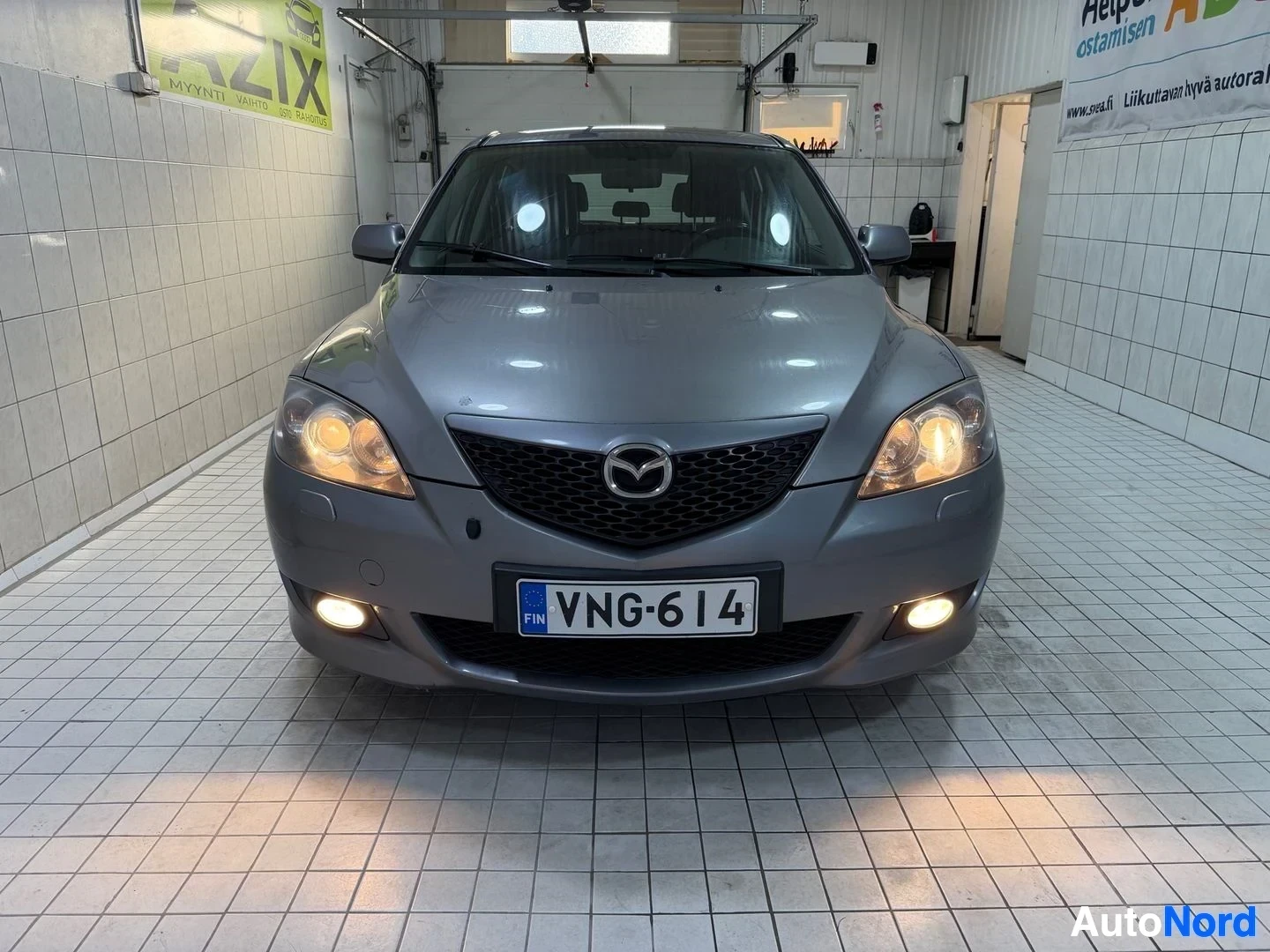 Edestä: Harmaa Mazda-merkkinen henkilöauto katsottuna edestä, sisätiloissa lattiahallissa. Ajovalot syttyneet. Rekisteritunnus näkyvissä