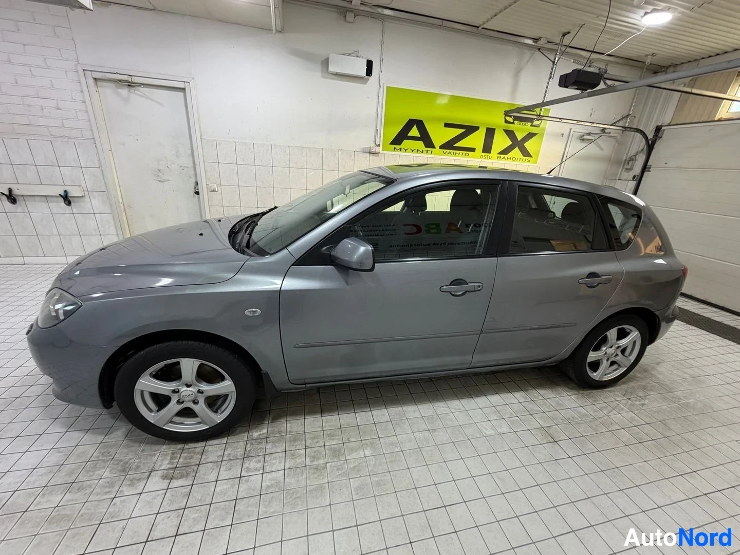 Sivulta: Hopeinen Mazda 3 henkilöauto, näkymä vasemmalta sivulta, oven avattuna, sisätiloissa.