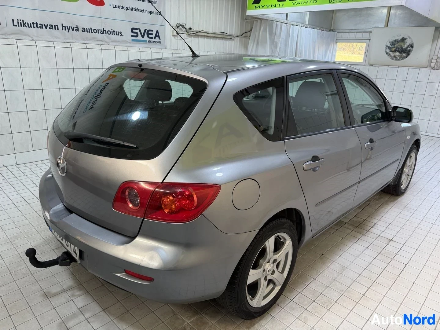 Takaviisto: Hopeinen Mazda 3 henkilöauto, näkymä takaa oikealta puolelta kolmen neljänneksen kulmasta, sisätiloissa.