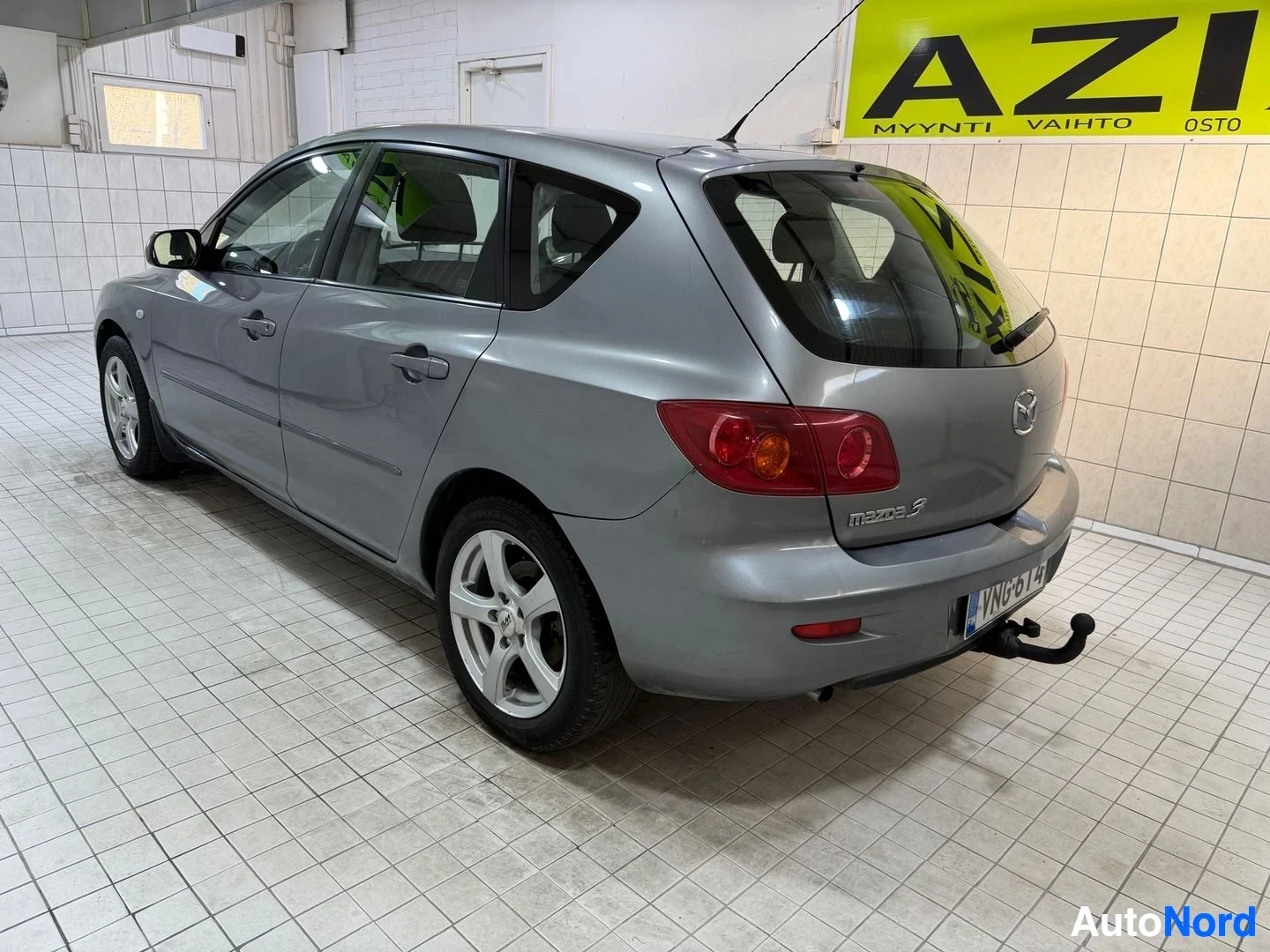 Takaviisto: Harmaa Mazda 3 sivusta takatilassa, näkyy oikea sivu ja taka, rekisterikilvessä näkyy VN64L, valkoinen vanteet, mustan väriset takavalo