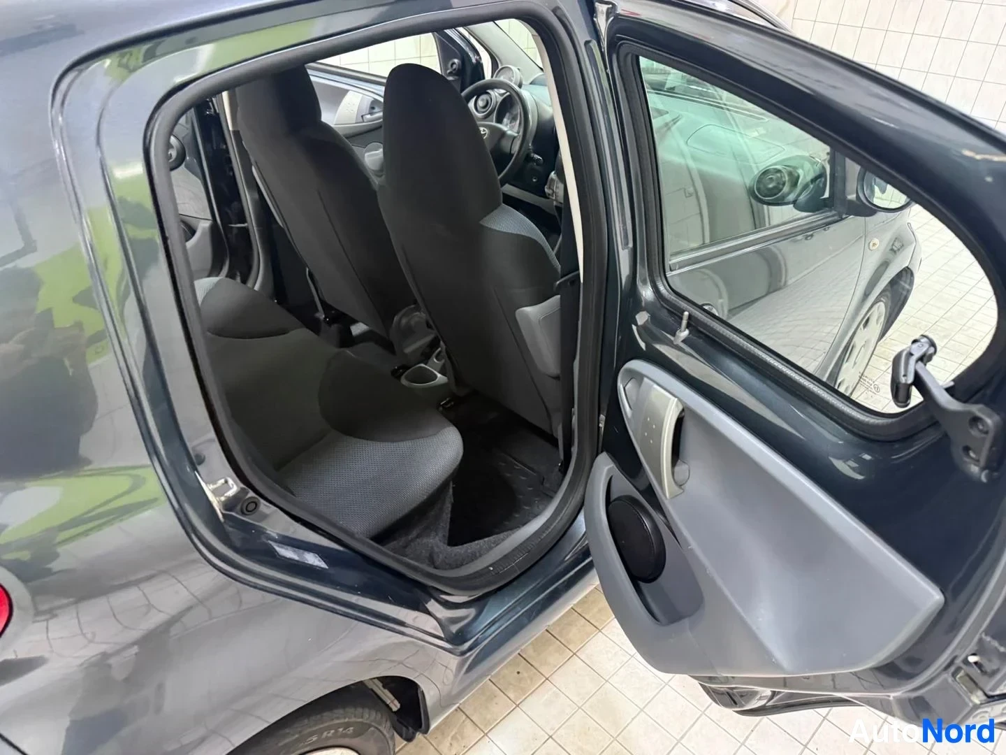 Ohjaamo: Avonainen auto, musta istuinvuori ja harmaa sisustus, avoin kuljettajan puoleinen ovi, näkyy ohjauspyörä ja kojelauta