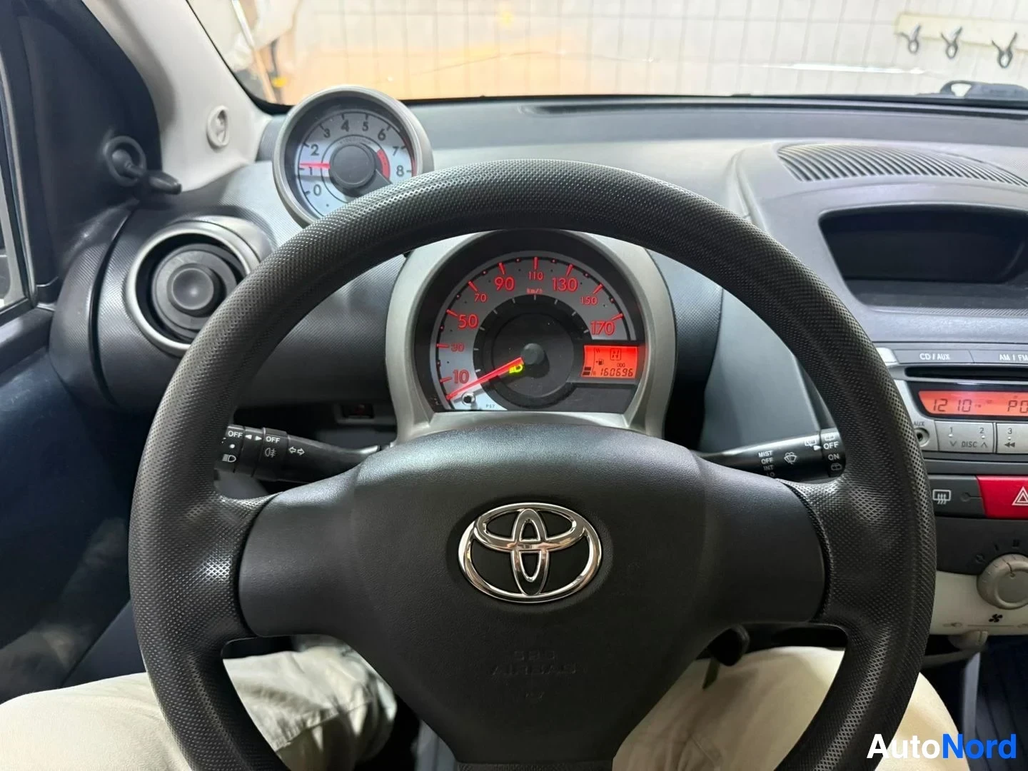 Yksityiskohta: Lähikuva mittarista ja ratista, punainen nelineulainen nopeusmittari ja Toyota-logo näkyvissä.