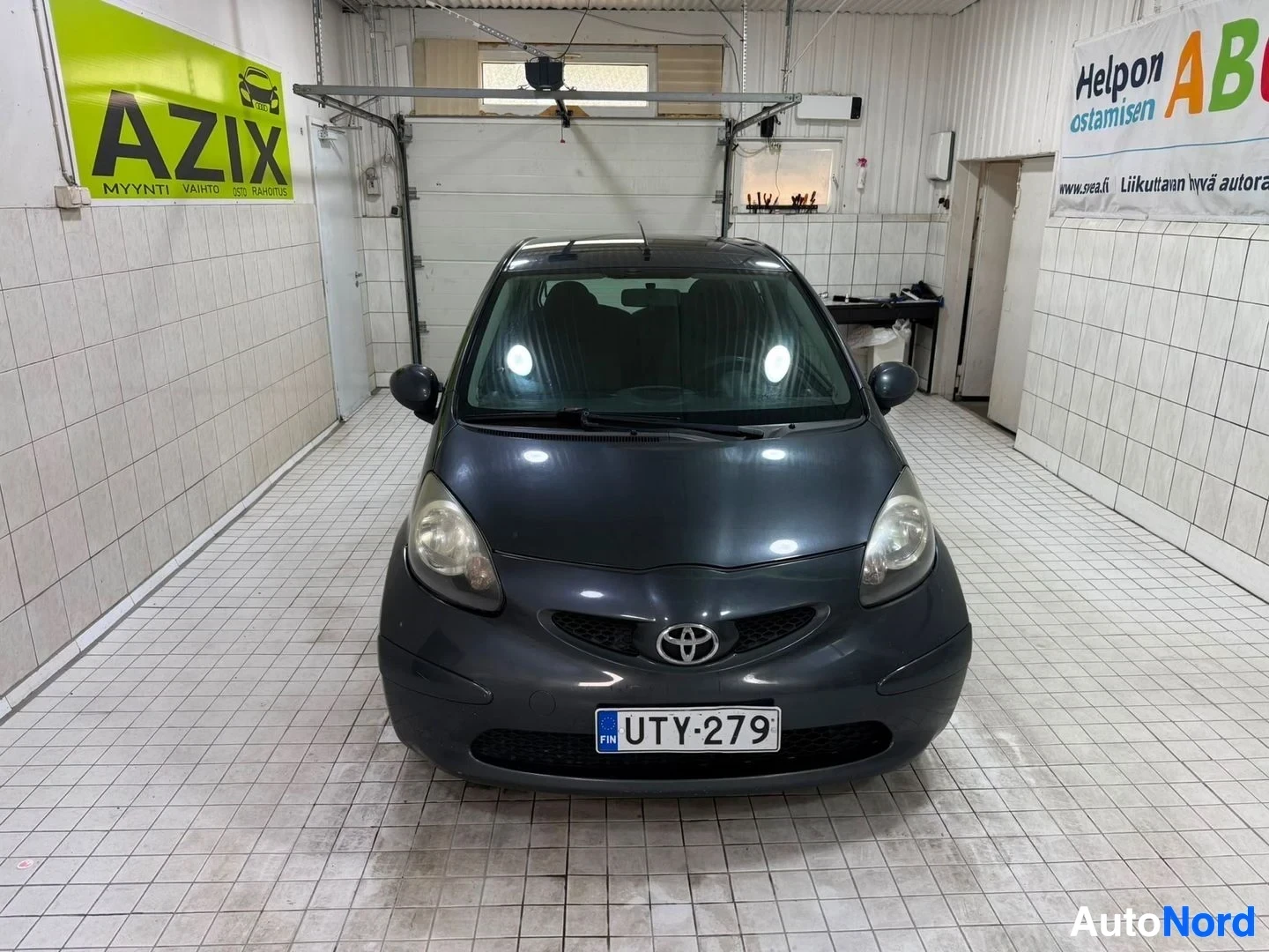 Edestä: Harmaa Toyota Aygo etulta katsottuna sisätiloissa, rekisterikyltti näkyvissä