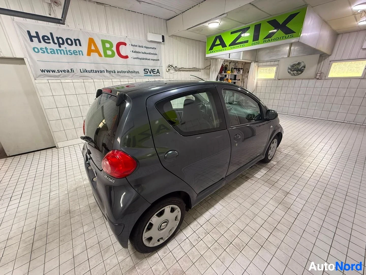 Sivulta: Tummansininen Toyota Aygo, kuvattu sisätilassa vasemmalta sivulta. Ovi on avoimena. Näkyy P&P Autohuo!to Ky:n palvelun merkintöjä seinällä.