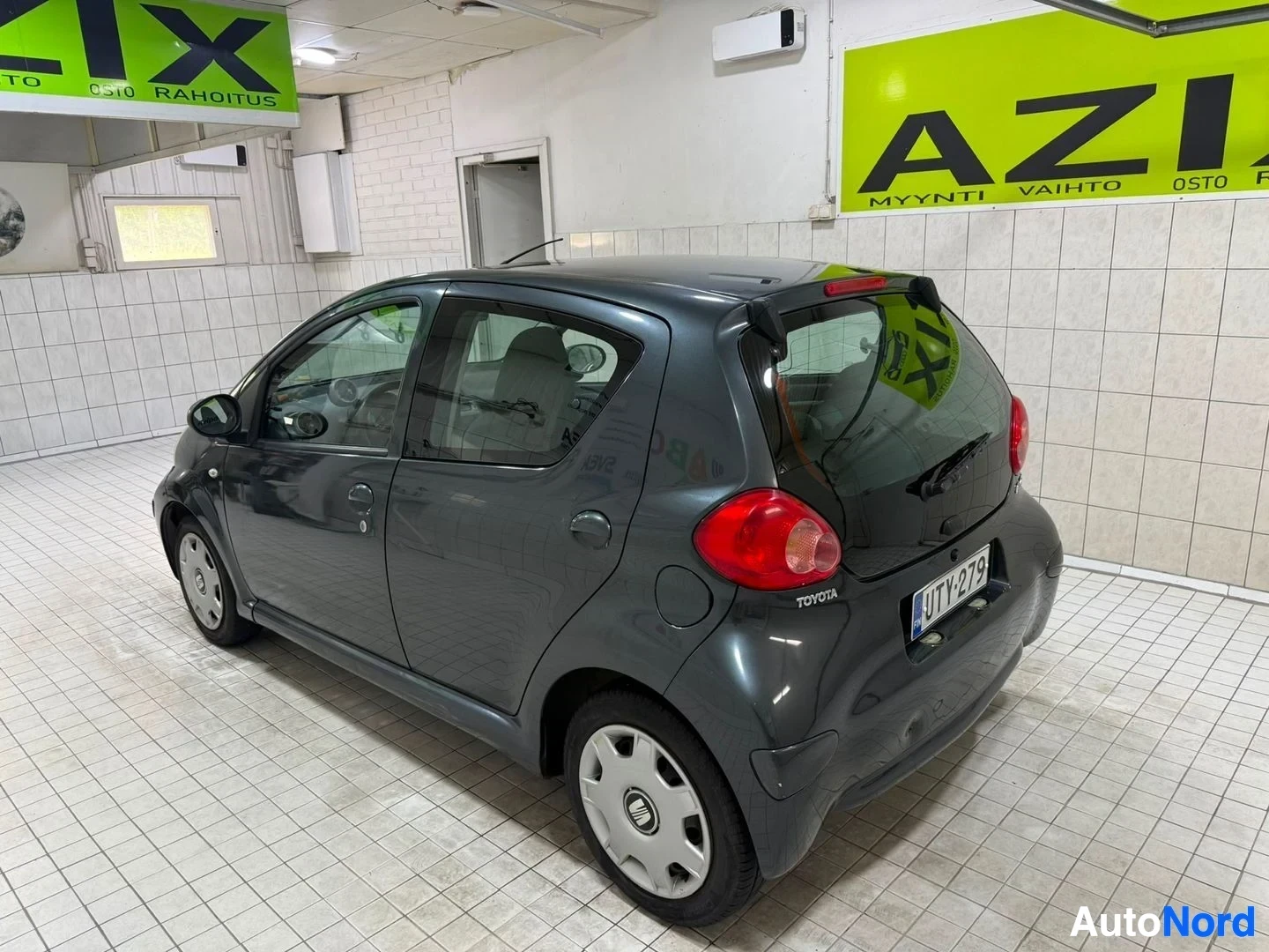 Takaviisto: Harmaa Toyota Aygo, kuvattu oikealta sivulta ja taakaa, ovi auki, näkyy takavalot ja rekisterikilpi