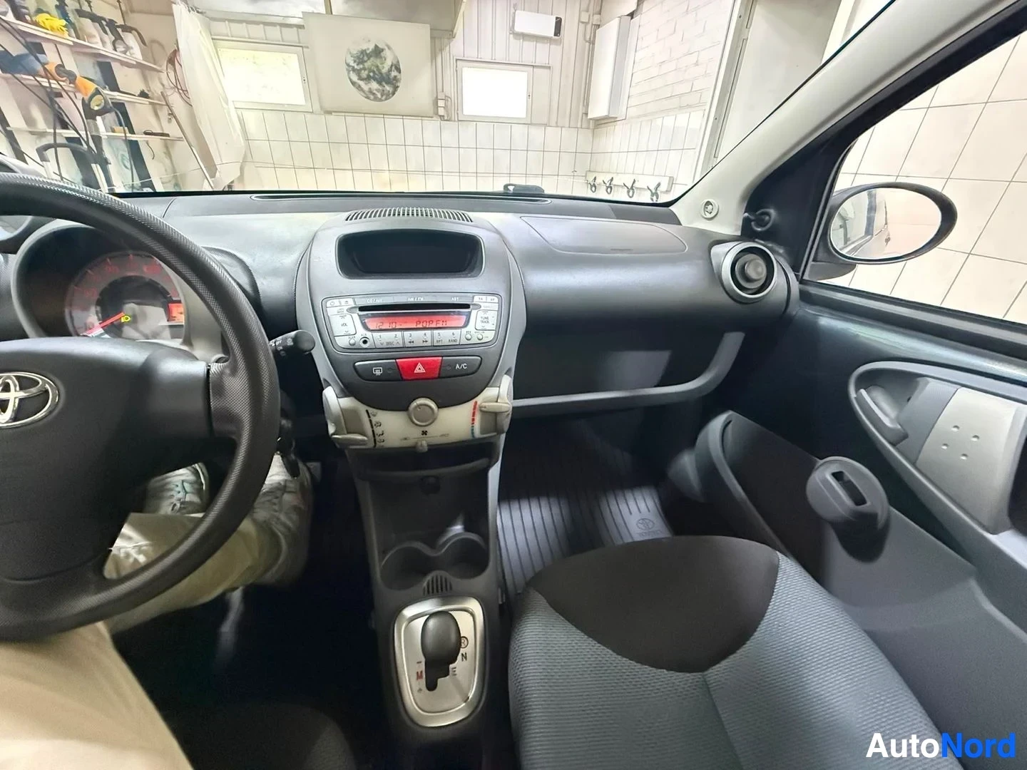 Ohjaamo: Auton sisustanäkymä etupenkiltä. Näkyy Toyota-ohjauspyörä, värilliset mittarit, radioyksikkö keskikonsolin päällä, ilmastointi-painikkeilla varustettu paneeli,