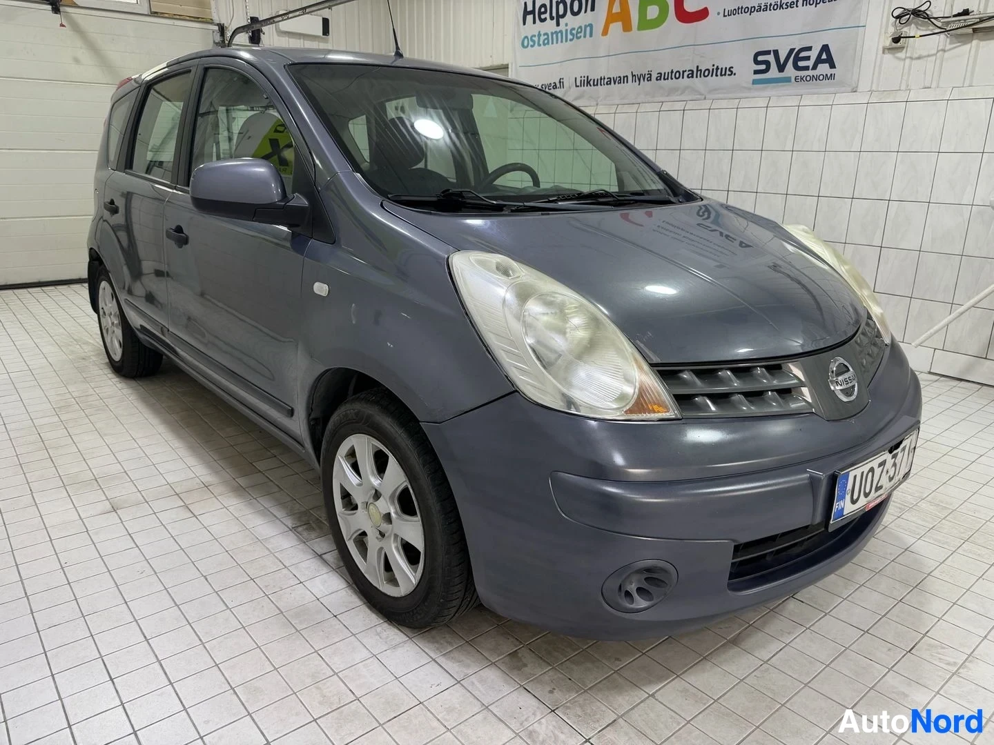 Etuviisto, nokka oikealle: Harmaa Nissan Note, kuvattu tasosta edessä vasemmalla. Rekisterikilvessä näkyy "UO7257T". Taustalla näkyy seinä ja palvelupiste "Helpon ostamisen ABC" -l