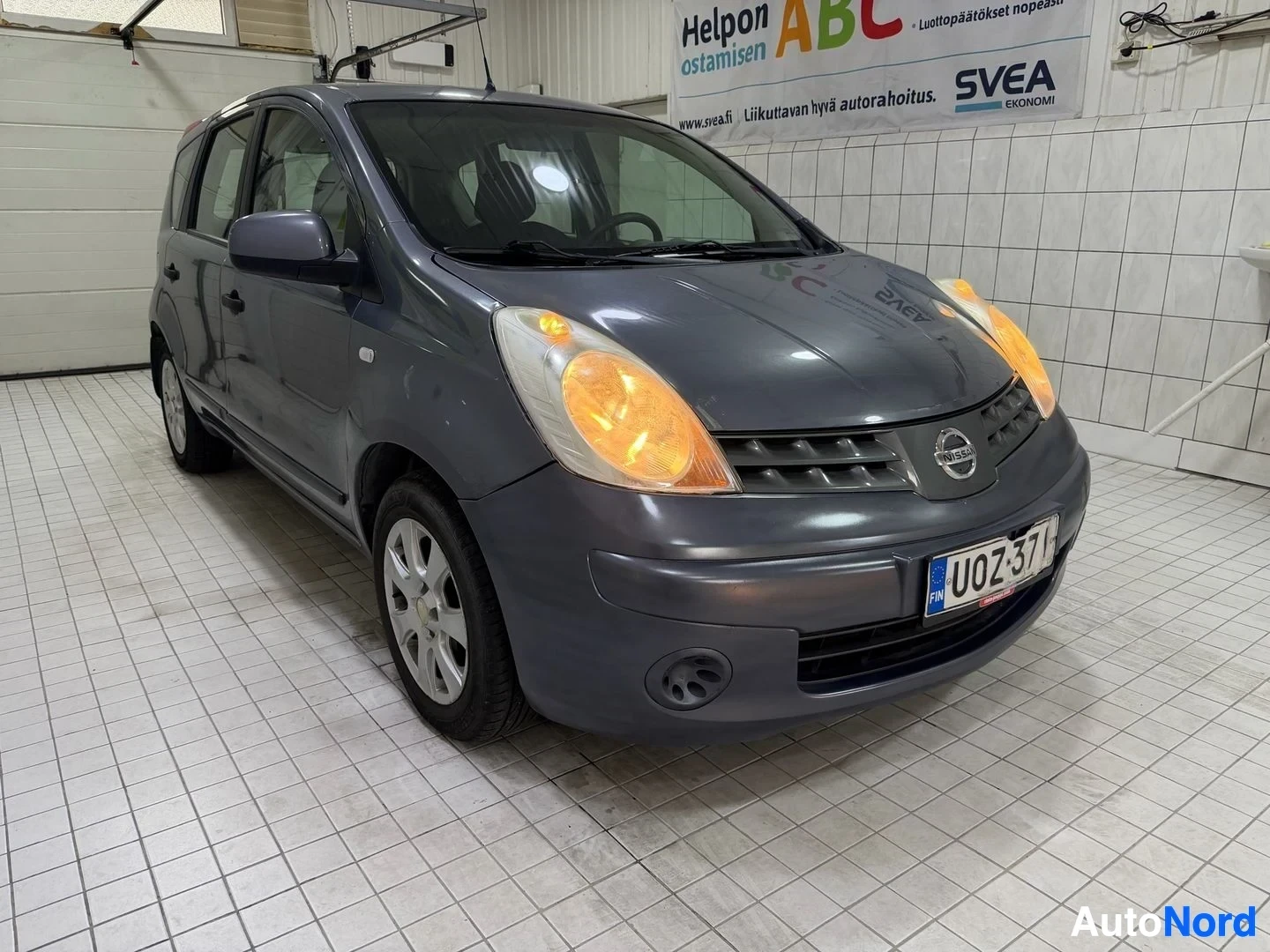Etuviisto, nokka vasemmalle: Nissan Note -kuntokuljetus tallin sisällä. Tummansinisen värin ajoneuvo kuvattuna vasemmalta sivulta. Rekisterikilvessä näkyvät tunnukset. Oran
