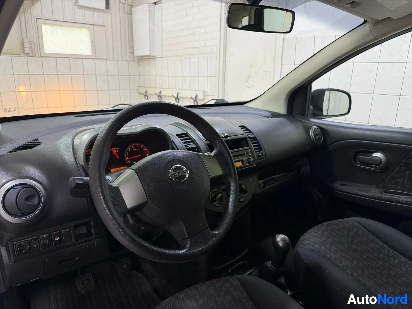 Ohjaamo: Kuljettajan asento sisältä: musta ohjausratas Nissan-logolla, puna-oranssi mittarit, musta paneeli, tummat kangaskudokset istuimissa, peiliö sisäkattoon.