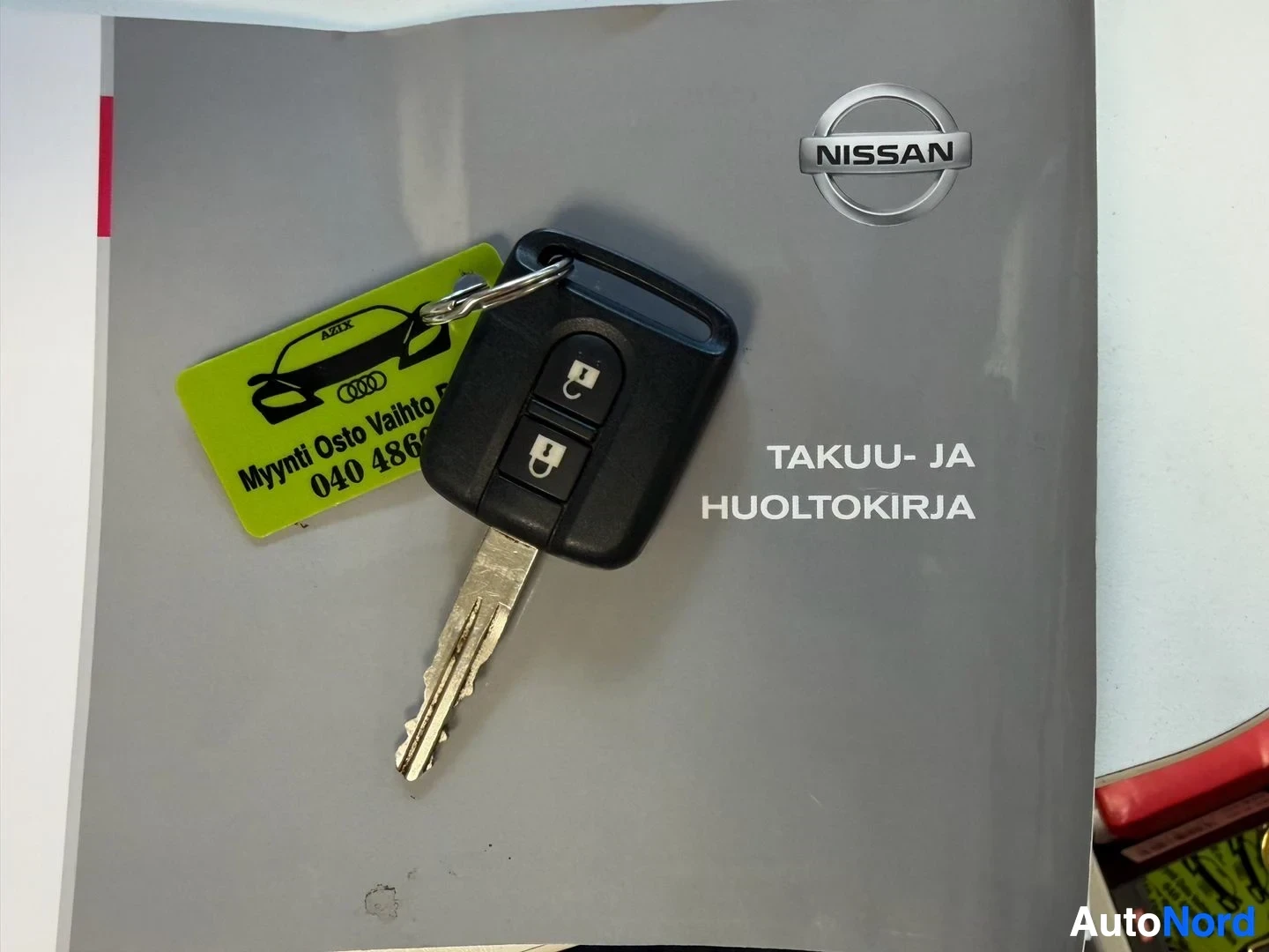 Asiakirja: Nissan avain mustalla kumitaitteella, jossa kaksi nappia, sekä keltainen varaston tunnistelappu ja Nissan-merkki harmaalla dokumentin taustalla. Teksti: TAKUU-