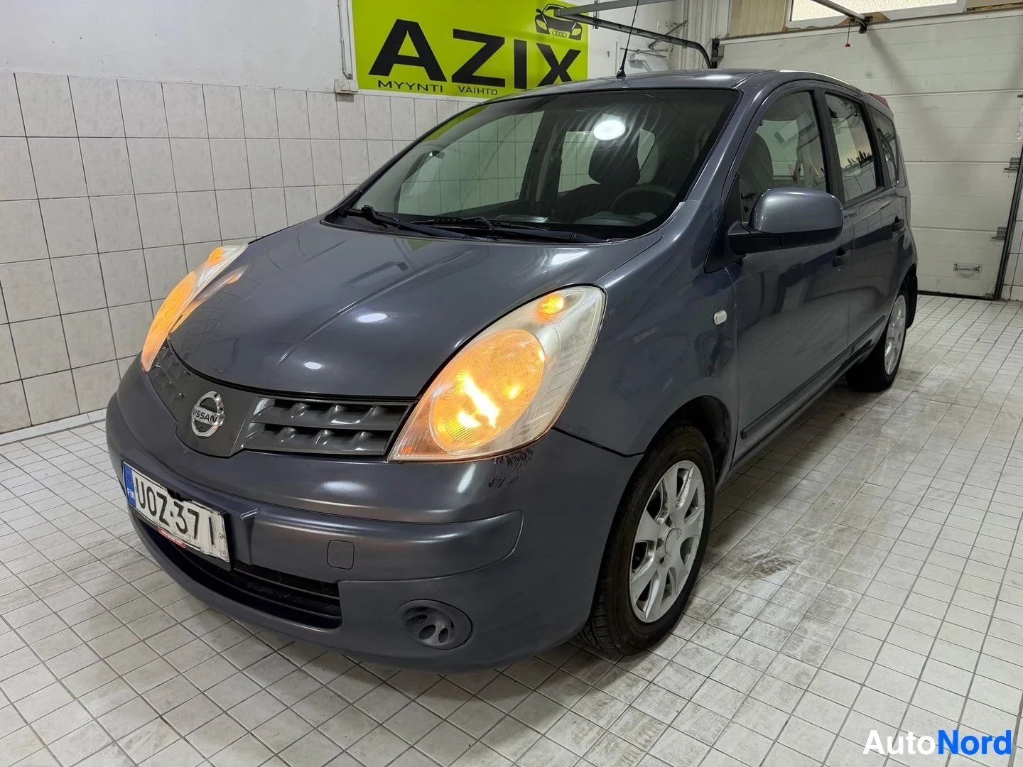 Etuviisto, nokka vasemmalle: Nissan Note -auto kuvattuna vasemmanpuoleisesta kulmasuunnasta hallissa. Tummansinisen värin ajoneuvo, oranssit ajovalot valaistu. Rekisterikilvessä UO