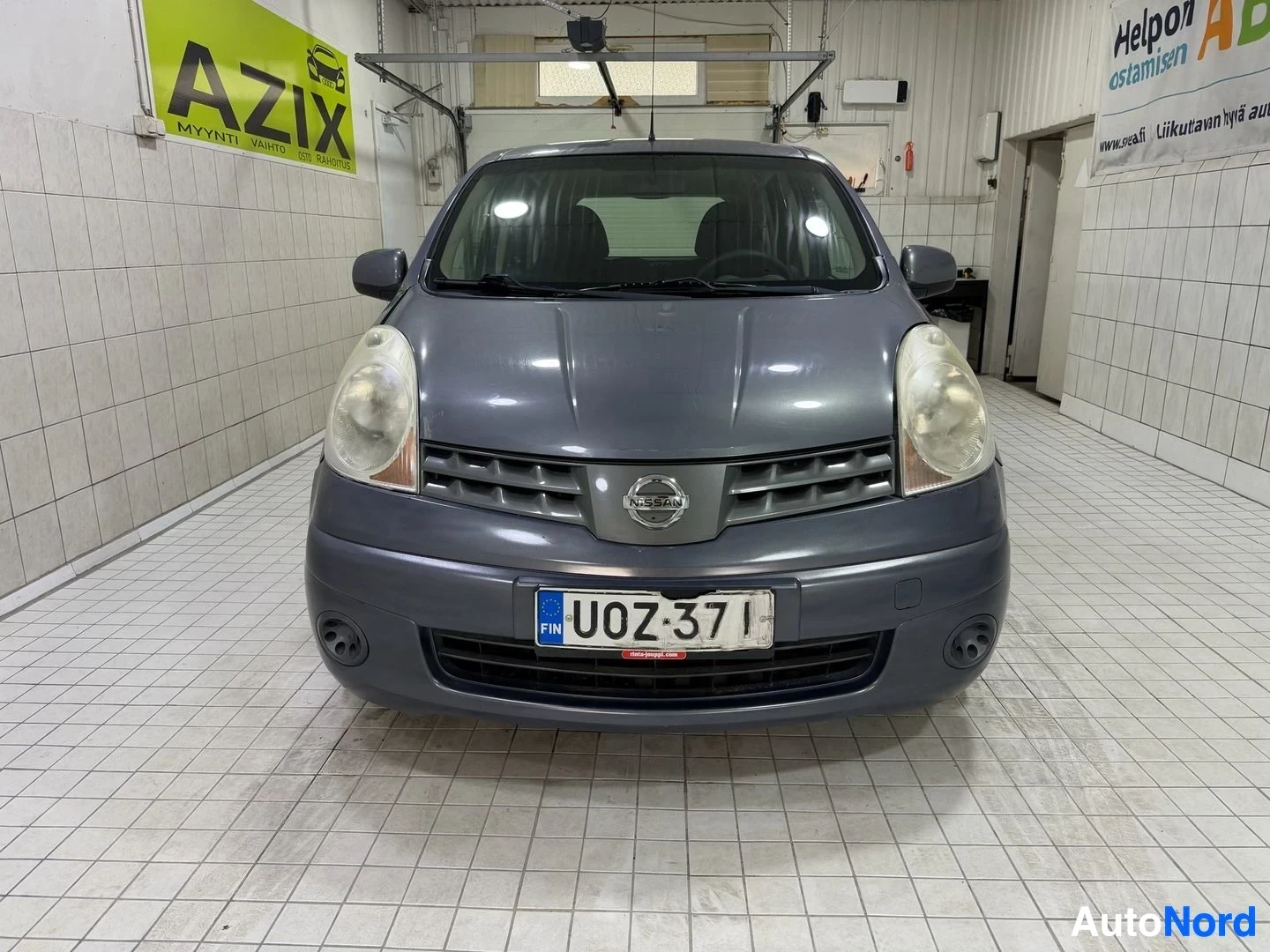 Edestä: Nissan Note edestä kuvattuna sisävarastossa. Rekisterikilvessä lukee. Harmaa auto, valkoisetksi muuttuneet ajovalot, mustat puskurit ja säleiköt