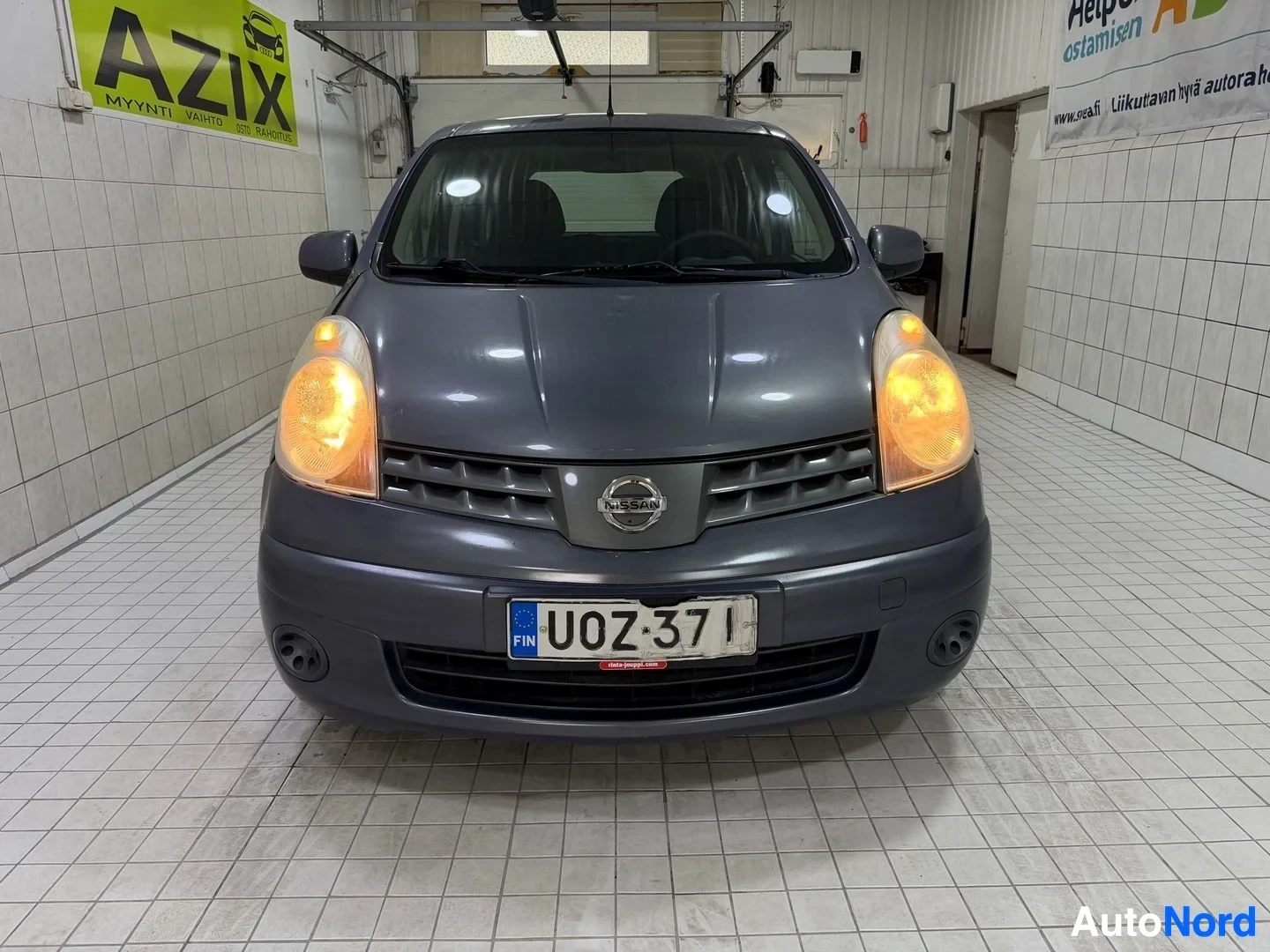 Edestä: Nissan Note -henkilöauto edestä päin katsottuna hallissa. Tummansinisen värin auto, oranssit ajovalot valaistu. Rekisterikilvessä. Valkoisia py öriä. Mu