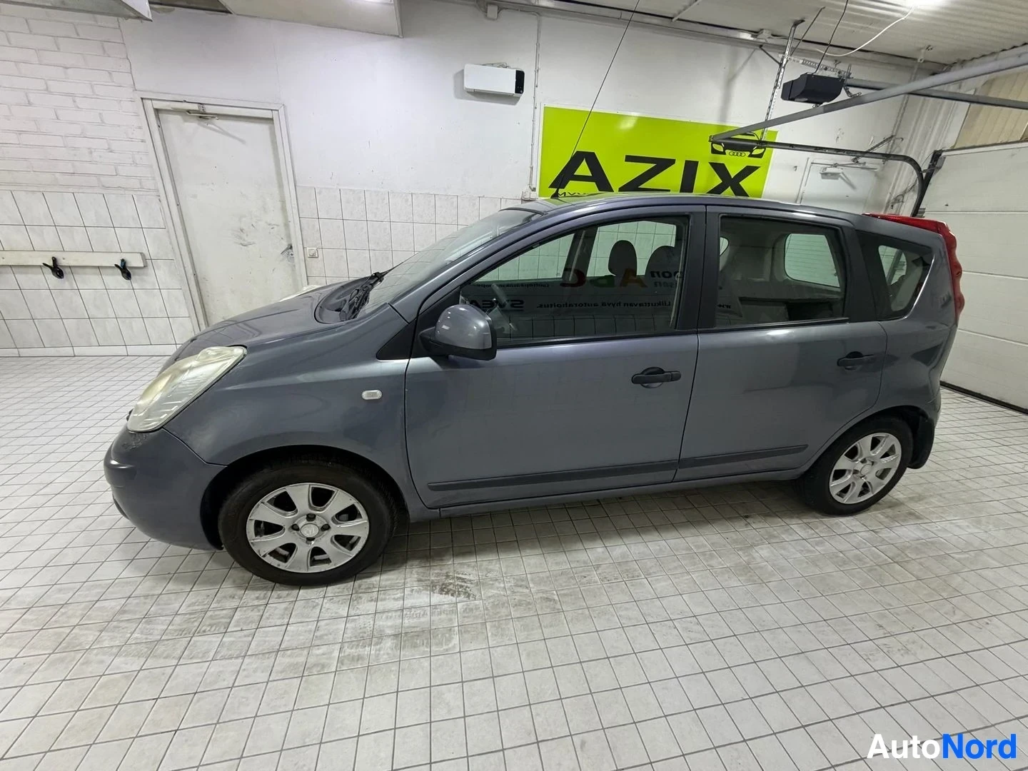 Sivulta: Harmaa Nissan Note, kuvattu sivulta oikealle päin. Auton ovi on auki. Taustalla näkyy AZIX-kyltti ja sisätilojen seinät.