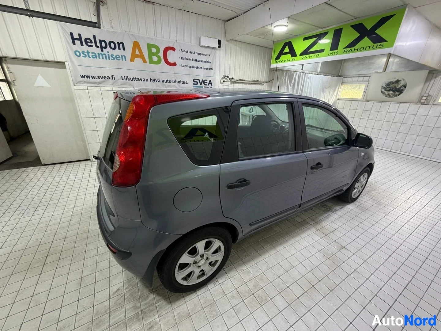 Sivulta: Harmaa Nissan Note pysäköitynä sisävarastoon, näkyy koko ajoneuvo sivusta. Punaiset takavalot, mustat ikkunat, hopeiset vantereet ja mustaksi maalattu katto.