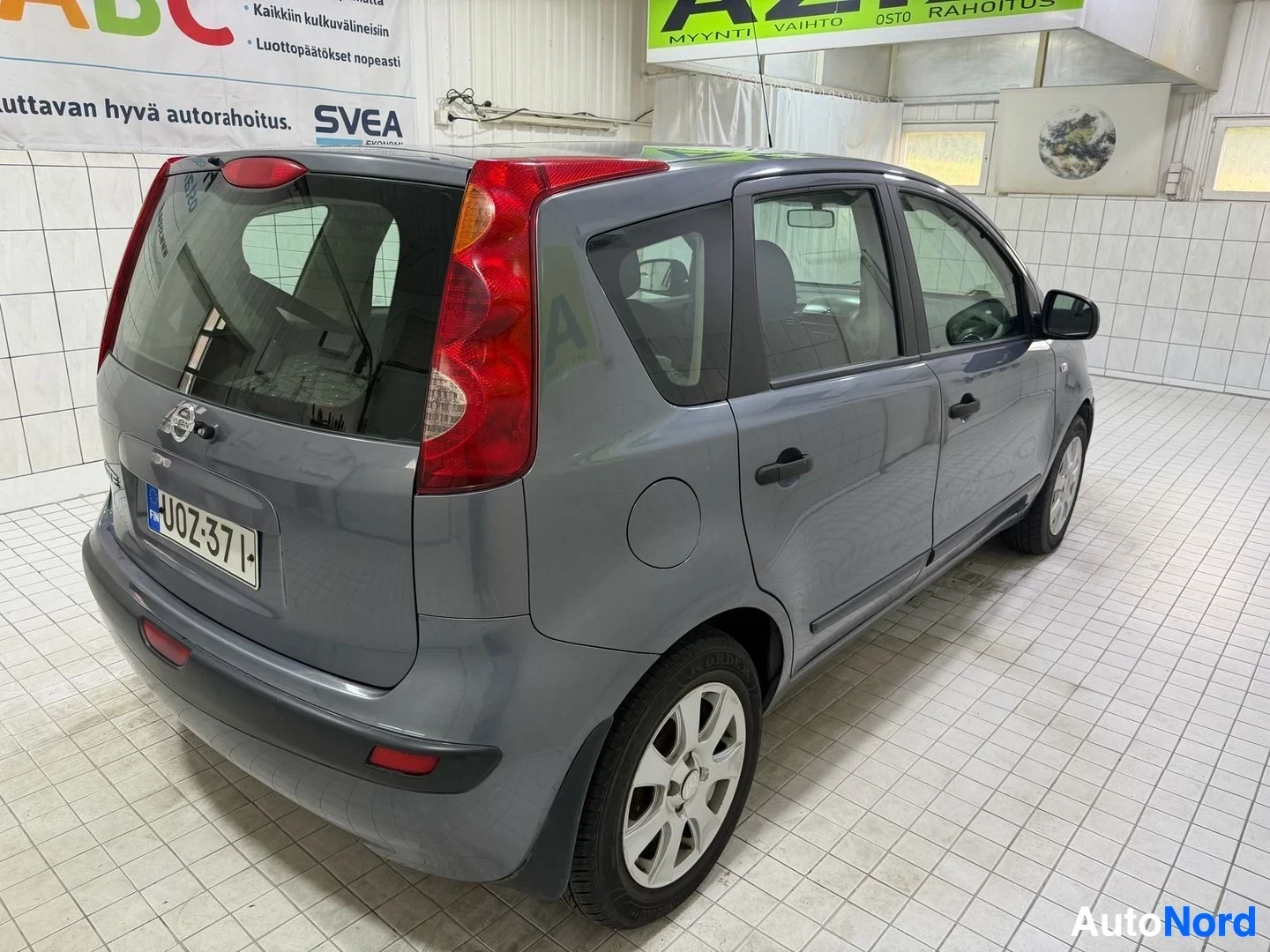 Takaviisto: Harmaa Nissan Note, kuvattu takaa kolmelta. Näkyy punainen takavalo ja harmaa takaovi. Rekisterikilvessä näkyy teksti "UO7257T". Taustalla näkyy sisätilojen sei