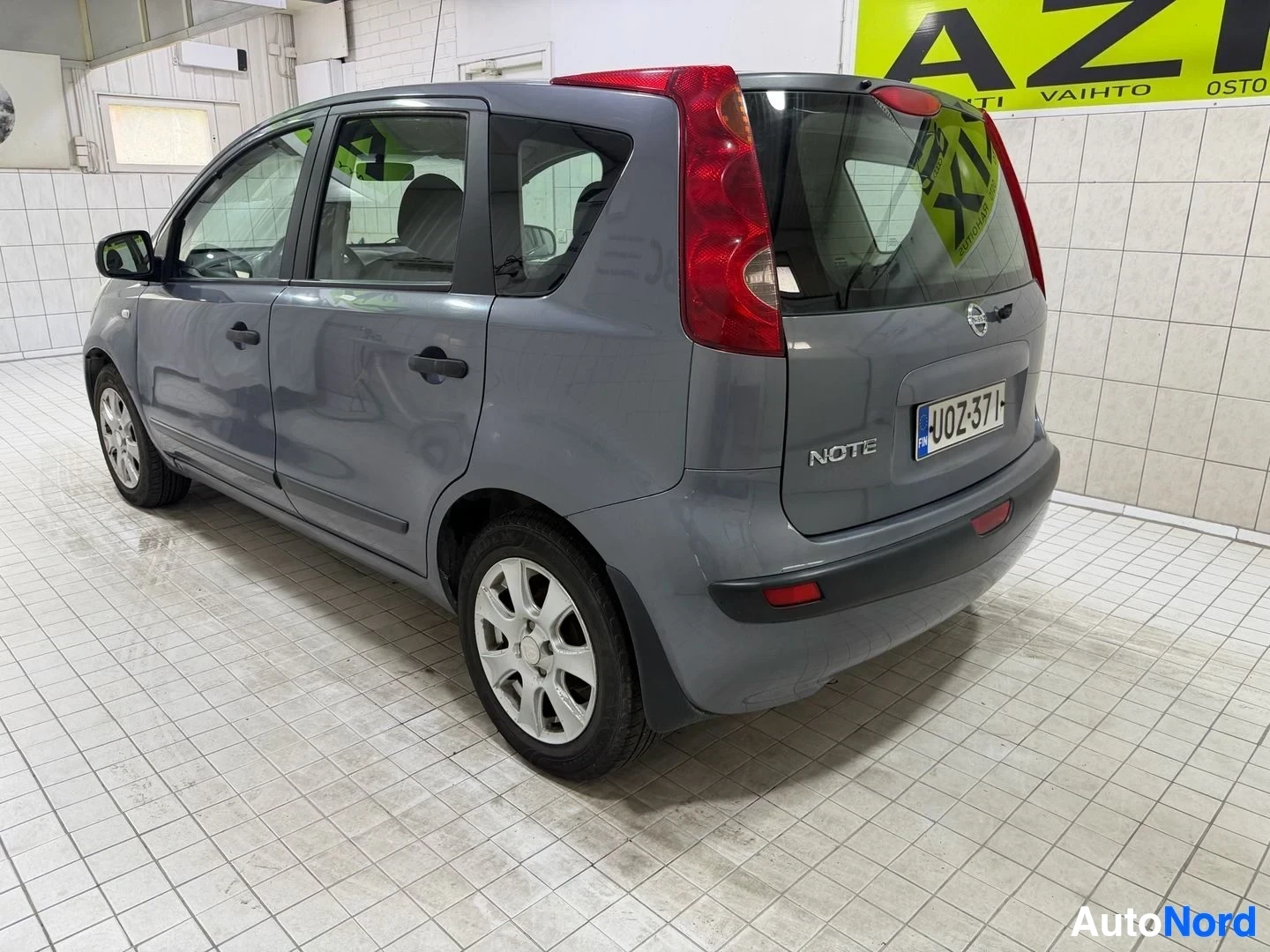 Takaviisto: Harmaa Nissan Note, oikealle suunnattu kolmen neljänneksen näkymä, vasen puoli näkyvissä, rekisterikilvessä JJO253TM, avattavat ovet, valkoinen seinä taustalla.