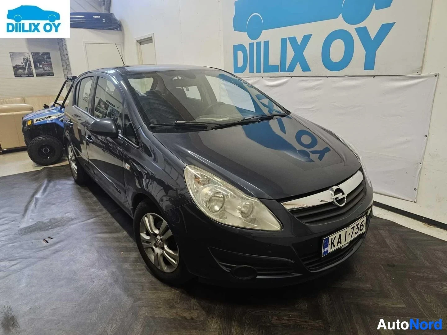 Etuviisto, nokka oikealle: Musta Opel Corsa -henkilöauto sisätiloissa, etupuoli ja vasen kylki näkyvät. Rekisterinumero KA-I-736. Ajovalot päällä.