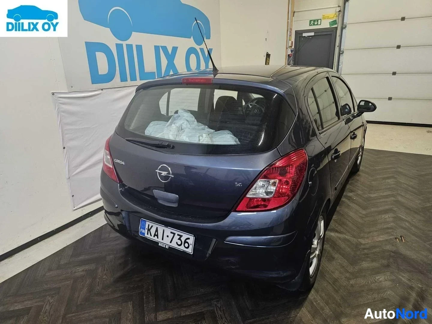 Takaviisto: Musta Opel Corsa -henkilöauto sisätiloissa, takapuoli ja oikea kylki näkyvät. Rekisterinumero KA-I-736. Takavalot punaiset. Korsa-teksti takaovet­ta.