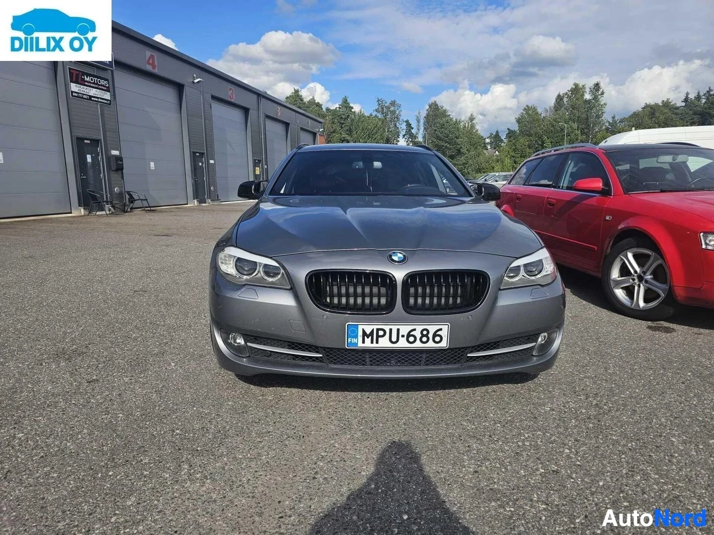 Edestä: Harmaa BMW-sedan etulta, näkyvillä rekisterikilvessä "". Taustalla punainen auto ja autoliikkeen rakennukset