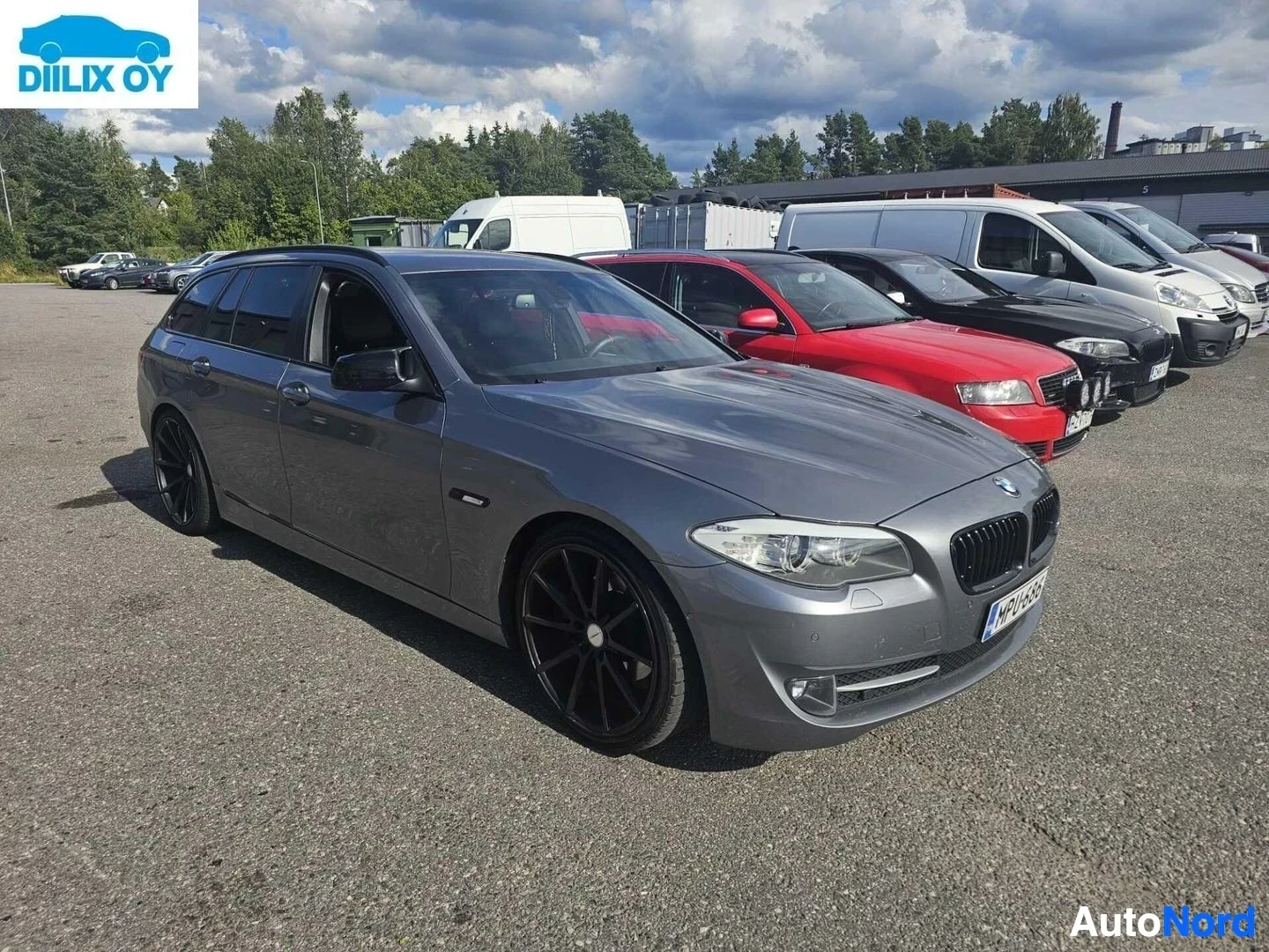 Etuviisto, nokka vasemmalle: Harmaa BMW-farmari parkkipaikalla muiden autojen kanssa, vasemmalle kääntynyt kolmen neljänneksen näkymä.