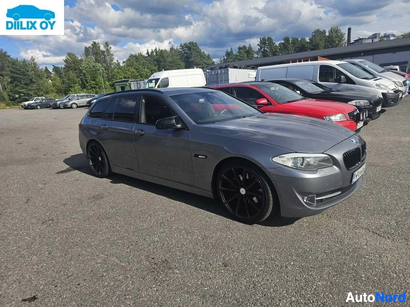 Etuviisto, nokka oikealle: Harmaa BMW-farmari pysäköitynä autoliikkeen pihalla, näkymä vasemmalta sivulta kolmelta neljäsosalta. Auto on suunnattu kohti kuvaa oikealle.