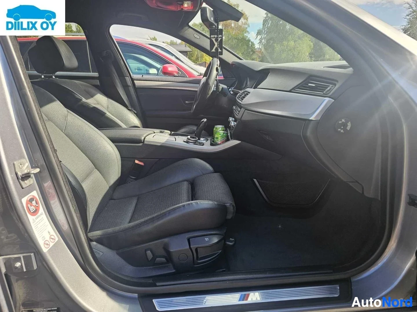 Etuistuimet: Auto sisäkuva, kuljettajan puoli. Näkyvät mustalla ja harmaalolla verhotellut istuimet, ohjauspyörä ja instrumenttopaneeli. Oven sisäpuolella näkyy M-logo.