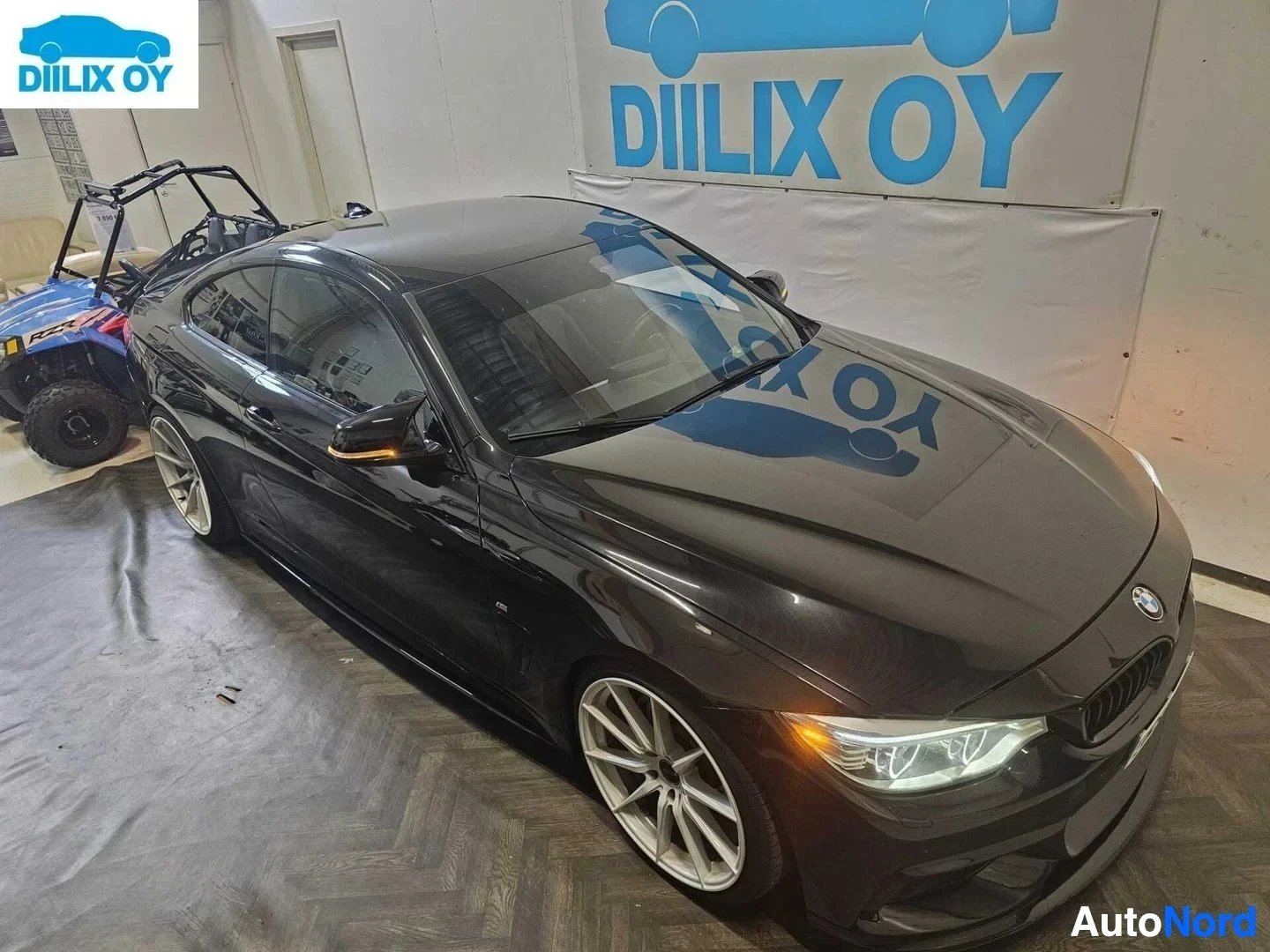 Etuviisto, nokka vasemmalle: Musta BMW coupe sisätiloissa, esillä oikeasta sivusta, valkoisen M-logolla varustetut vanteet näkyvissä
