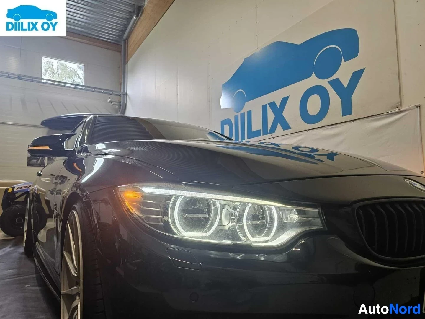 Sivulta: Musta BMW coupe vasemmalta sivulta, LED-ajovalot ja rengasmuotoiset silmät näkyvissä, harmaa tela taustalla