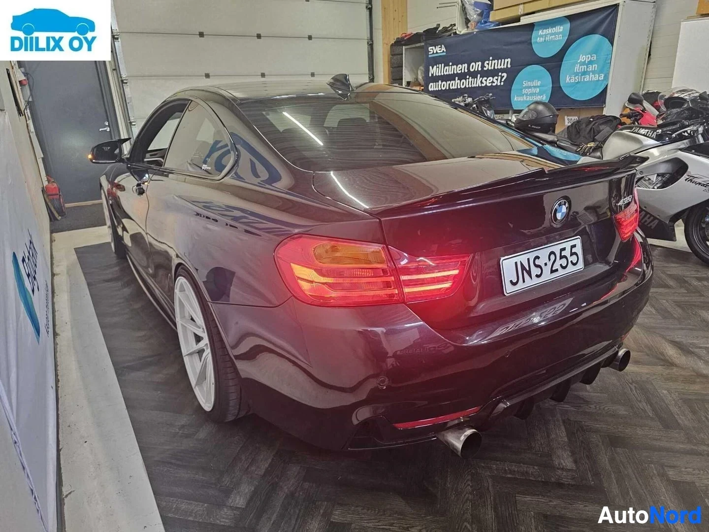 Takaviisto: Viininpunainen BMW-coupe sisätilassa, takaviistosta näkyvä oikea puoli, rekisterikilpi JNS 255 näkyvillä, LED-takavalo syttyneenä.