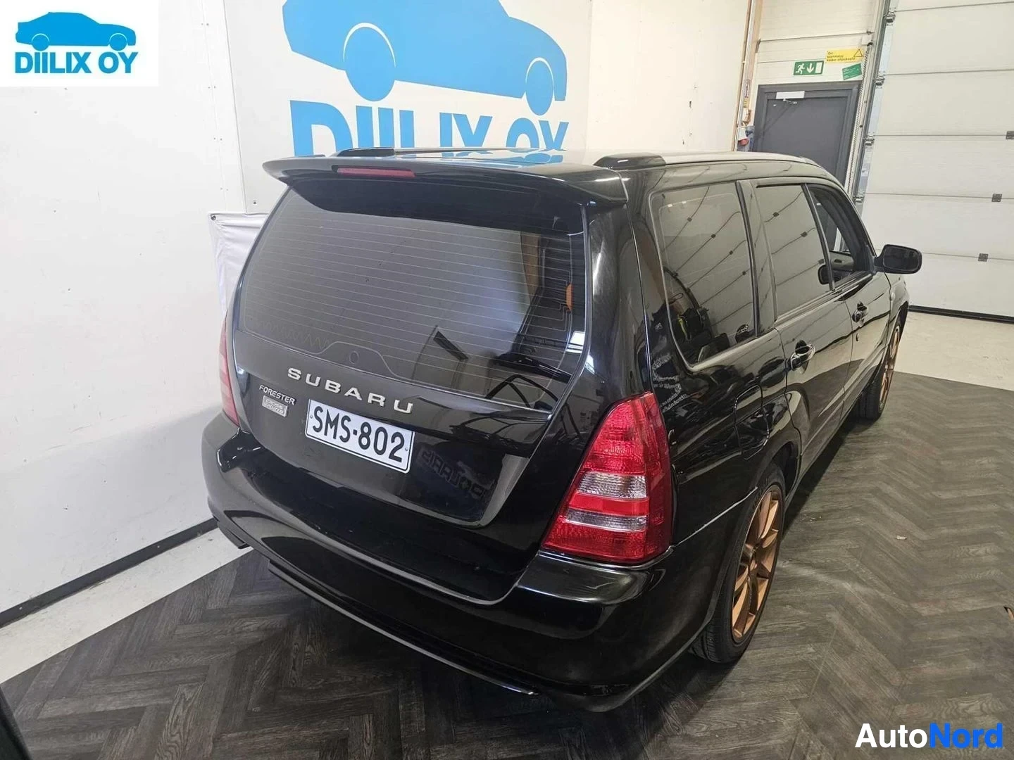 Takaa: Musta Subaru Forester -farmari sisätiloissa, kuvattu takaa. Näkyy rekisterikilvessä teksti ''. Ajoneuvolla punaiset takavalot ja pronssinväriset vanteet