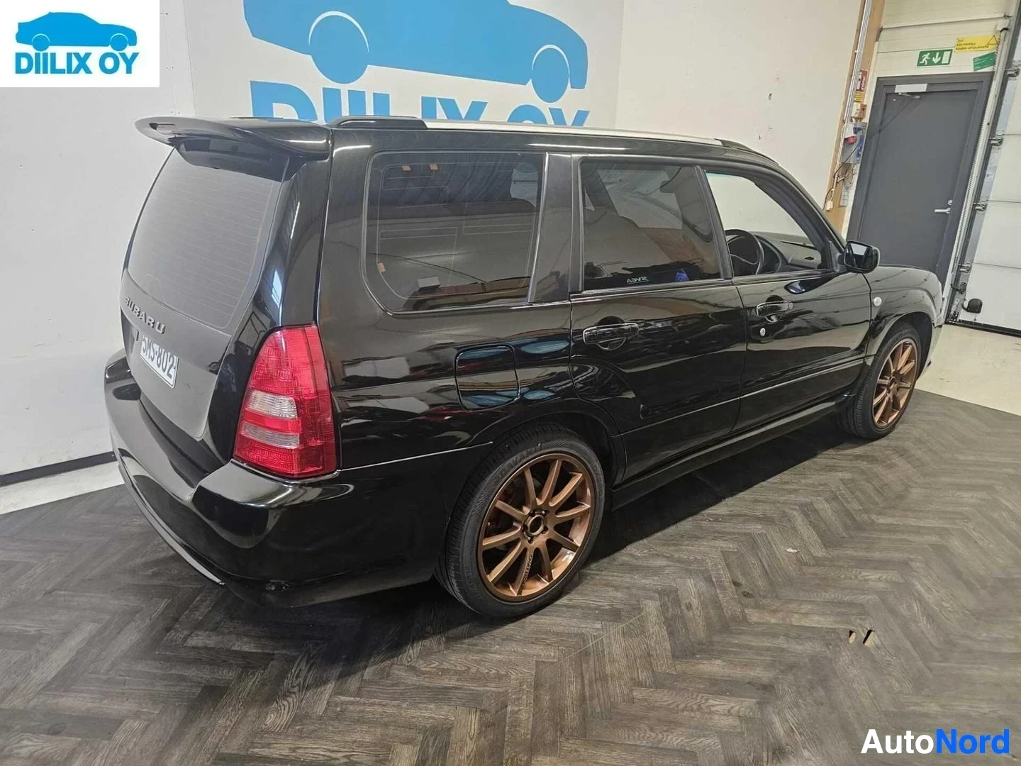 Takaviisto: Musta Subaru Forester -farmari sisätiloissa, kuvattu takakolmen neljänneksen kulmasta oikealta. Ajoneuvon takaa näkyy punaiset takavalot ja pronssinväriset vant