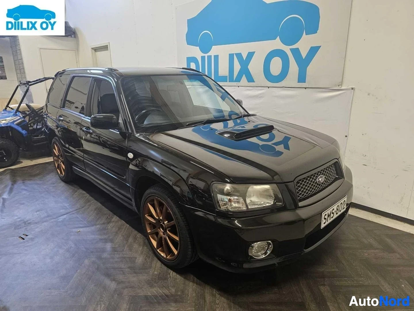 Etuviisto, nokka vasemmalle: Musta Subaru Forester kuvan vasemmalla puolella katsottuna sisätiloissa. Rekisteri. Oranssit vanteet. Avoin tavaratila vasemmalla