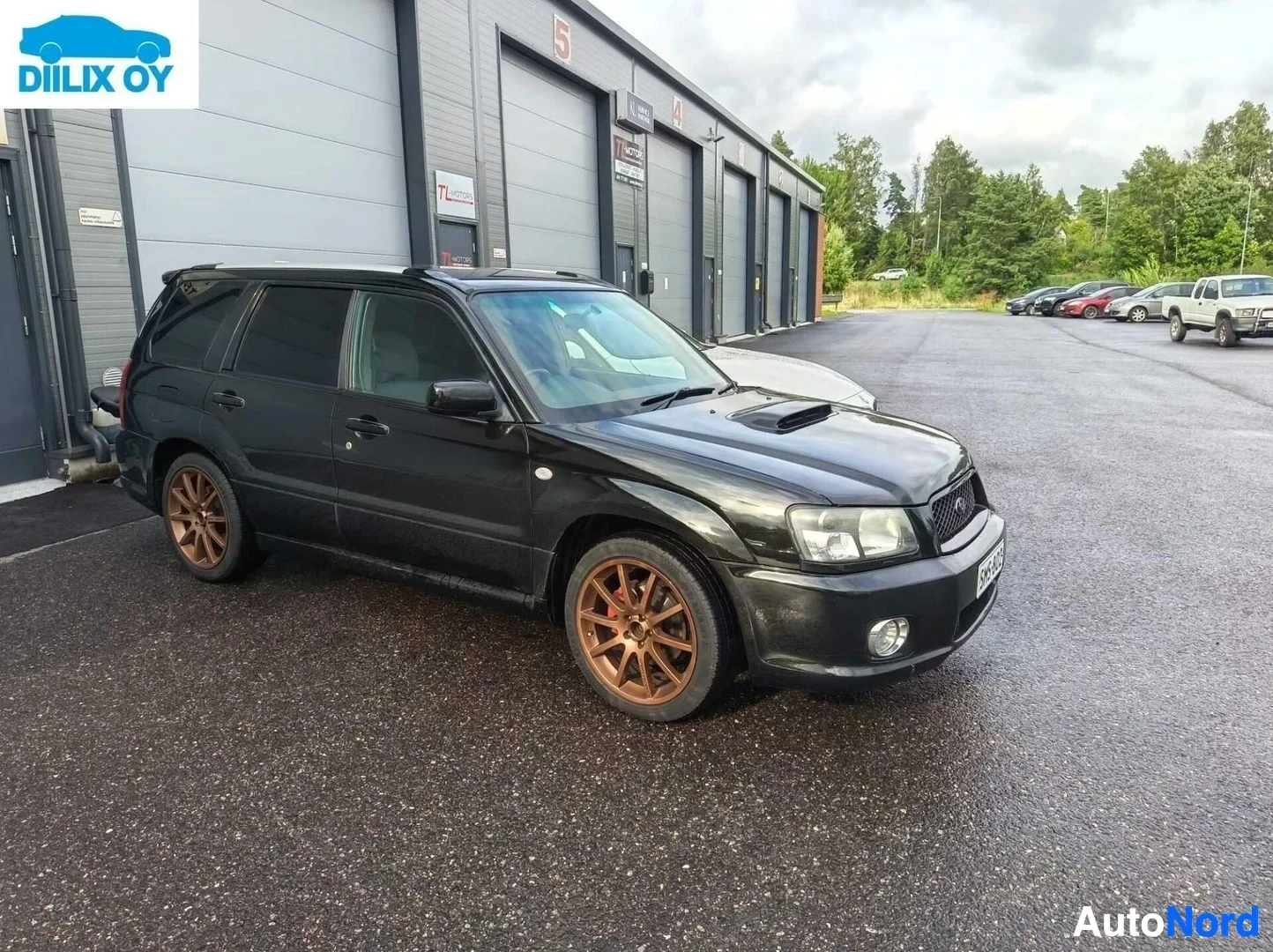 Etuviisto, nokka oikealle: Musta Subaru Forester, jossa on pronssit vanteet, pysäköitynä asfaltoitavalle alueelle varaston edessä.