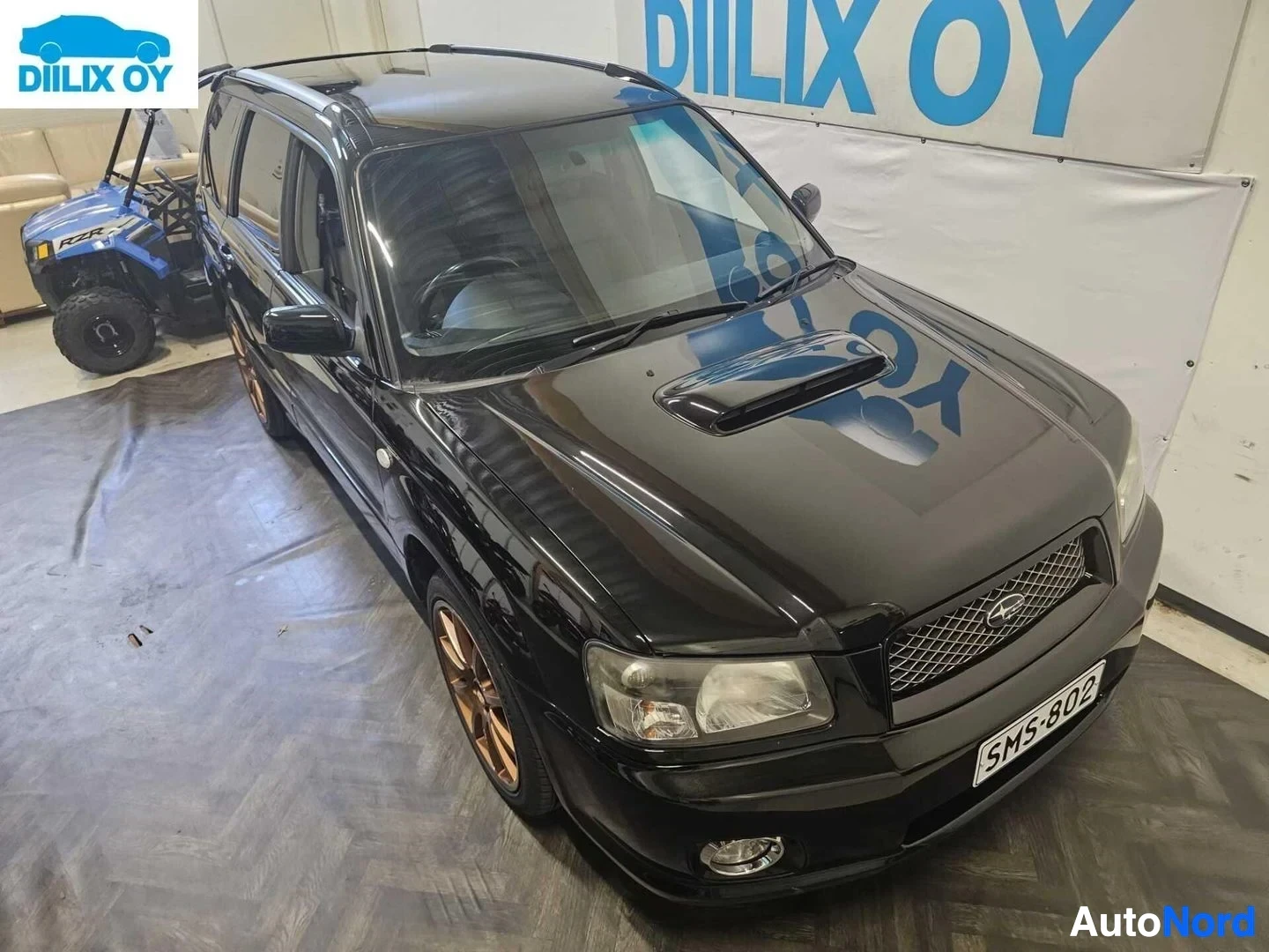 Etuviisto, nokka oikealle: Musta Subaru Forester kuvan oikealla puolella katsottuna sisätiloissa. Rekisteri. Oranssit vanteet. Moottoriventtiilit näkyvät konepellissa