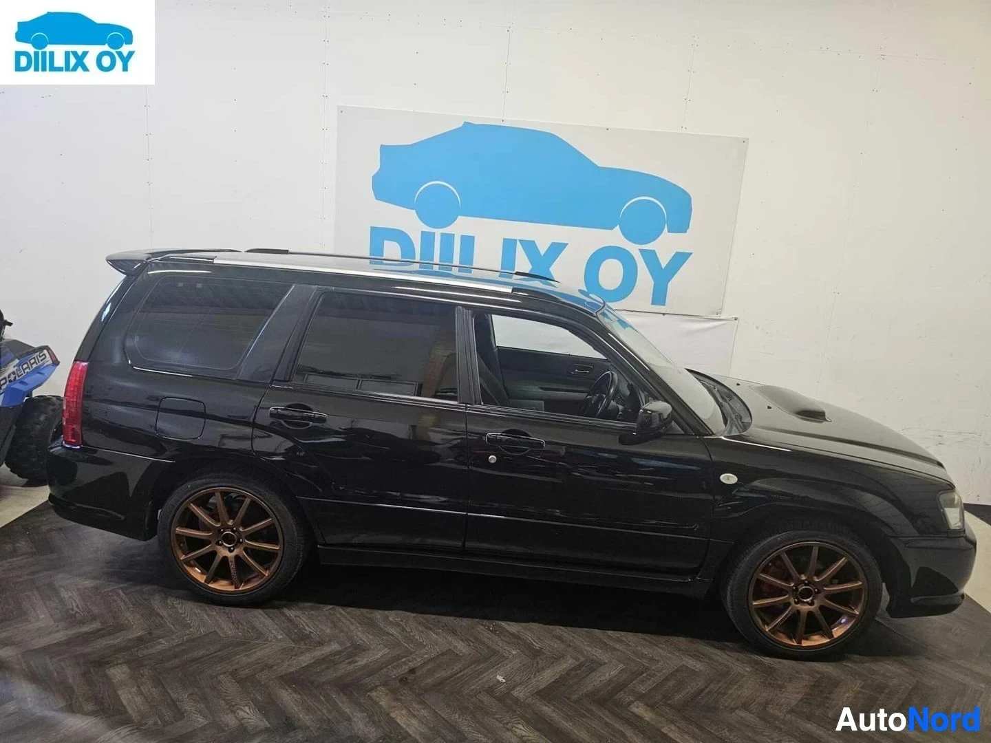 Sivulta: Musta Subaru Forester -farmari sisätiloissa, kuvattu sivusta vasemmalta. Ajoneuvolla on pronssinväriset vanteet ja punaiset takavalot. Taustalla näkyy sininen j