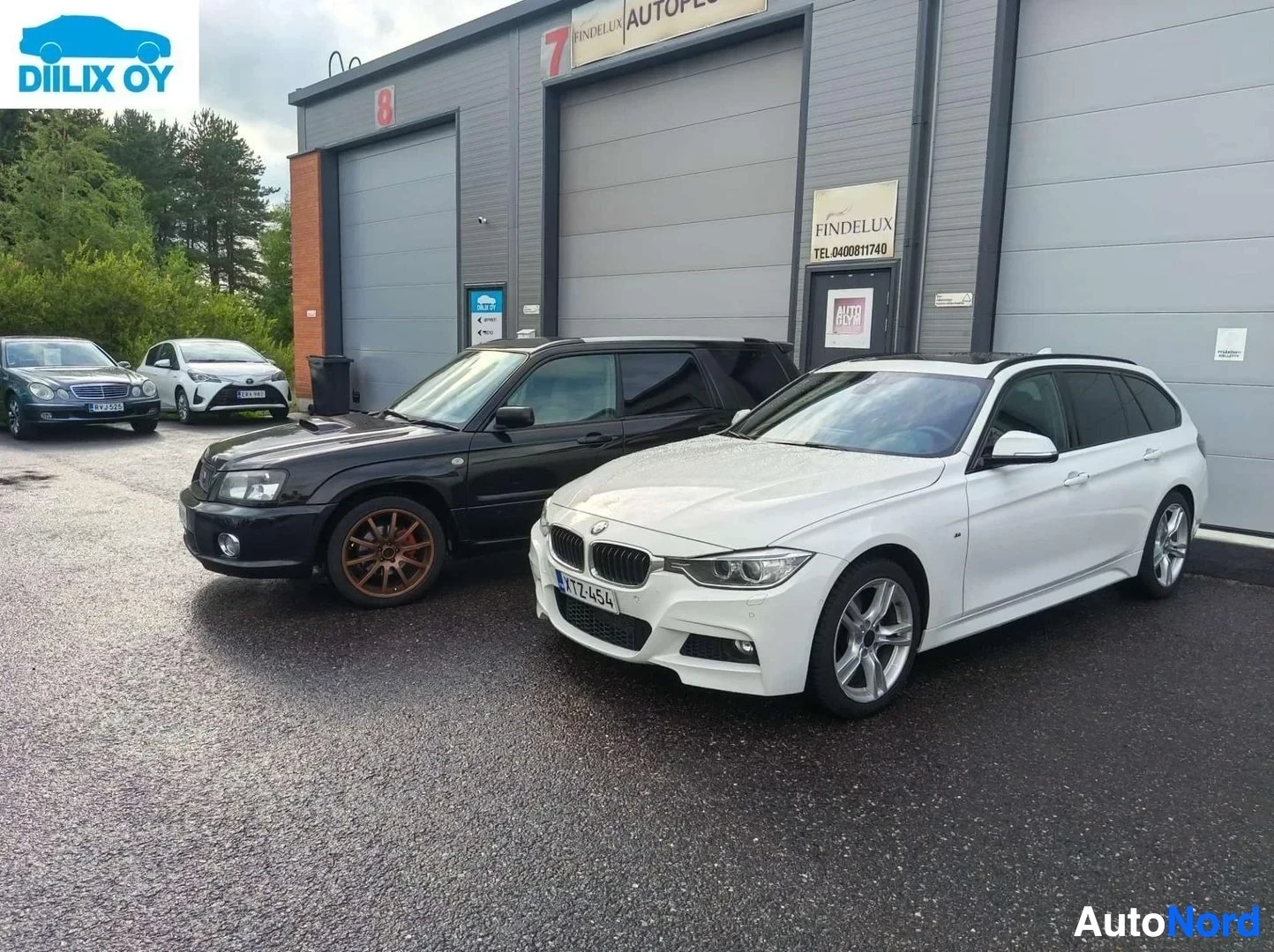 Sivulta: Musta Subaru Forester pronssisilla vanteilla ja valkoinen BMW seuraavansa rinnalla korjaamon edessä.
