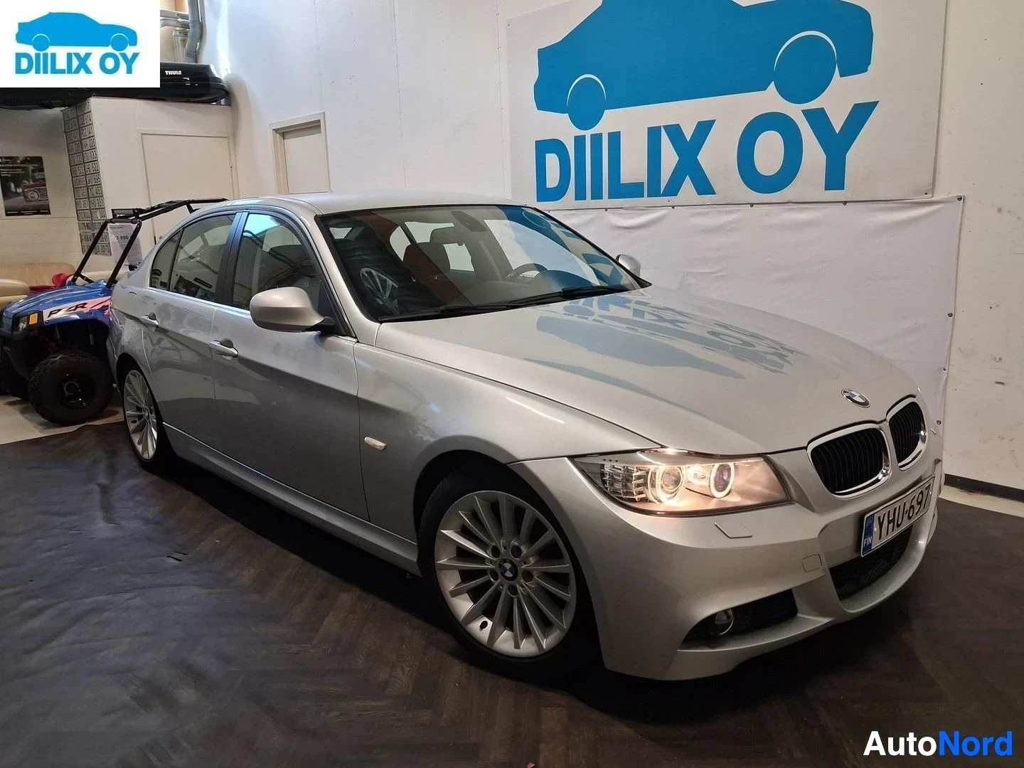 Etuviisto, nokka oikealle: Valkoinen BMW-sedan sivusta etupuoli kulmassa oikealle. Rekisterikilpi. LED-silmät valaistuina. Diilux Oy varaston tausta