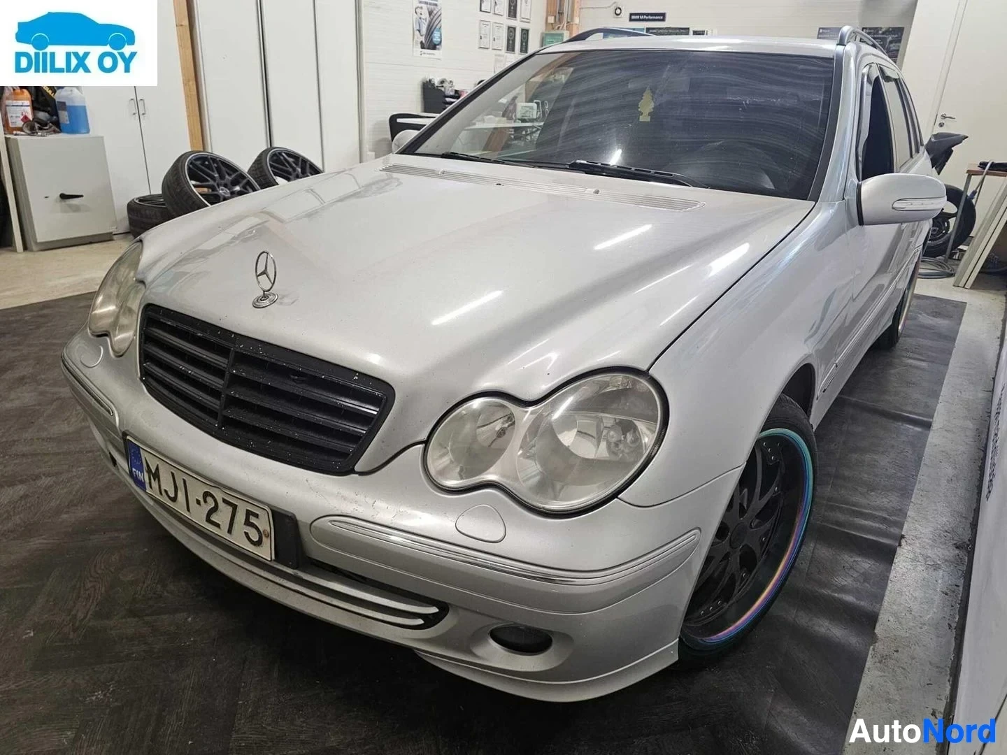 Etuviisto, nokka vasemmalle: Valkoinen Mercedes-Benz-farmari edestä oikealle, rekisterikilvessä, sisätiloissa