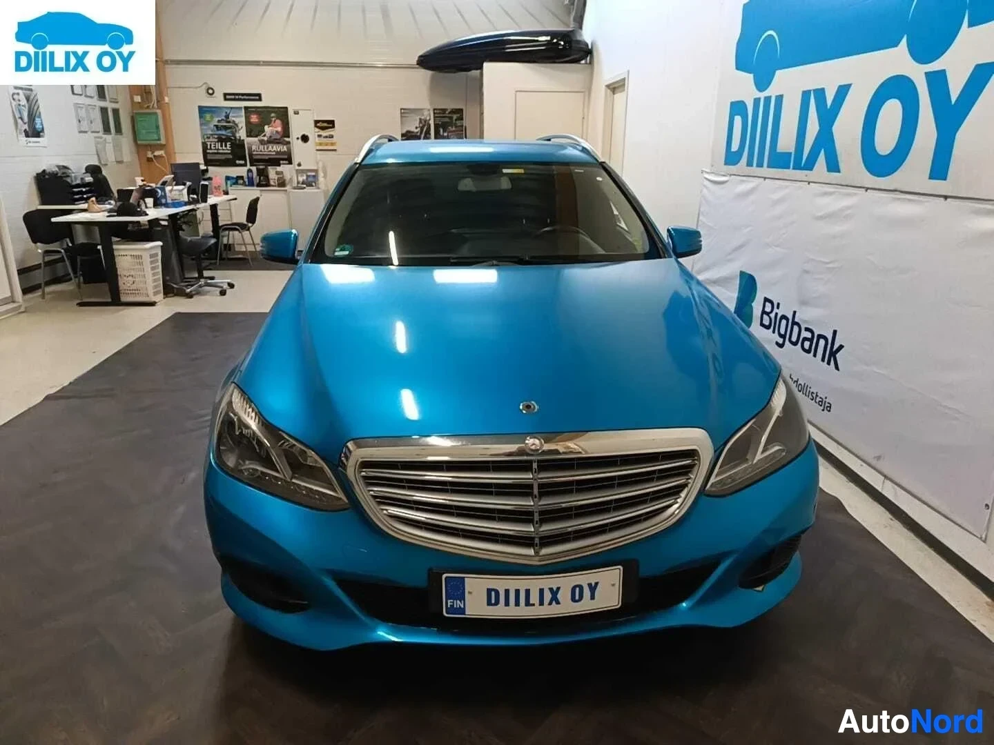 Edestä: Sininen Mercedes-Benz E-sarjan farmari varastossa edestä, rekisterikilpi näkyy.