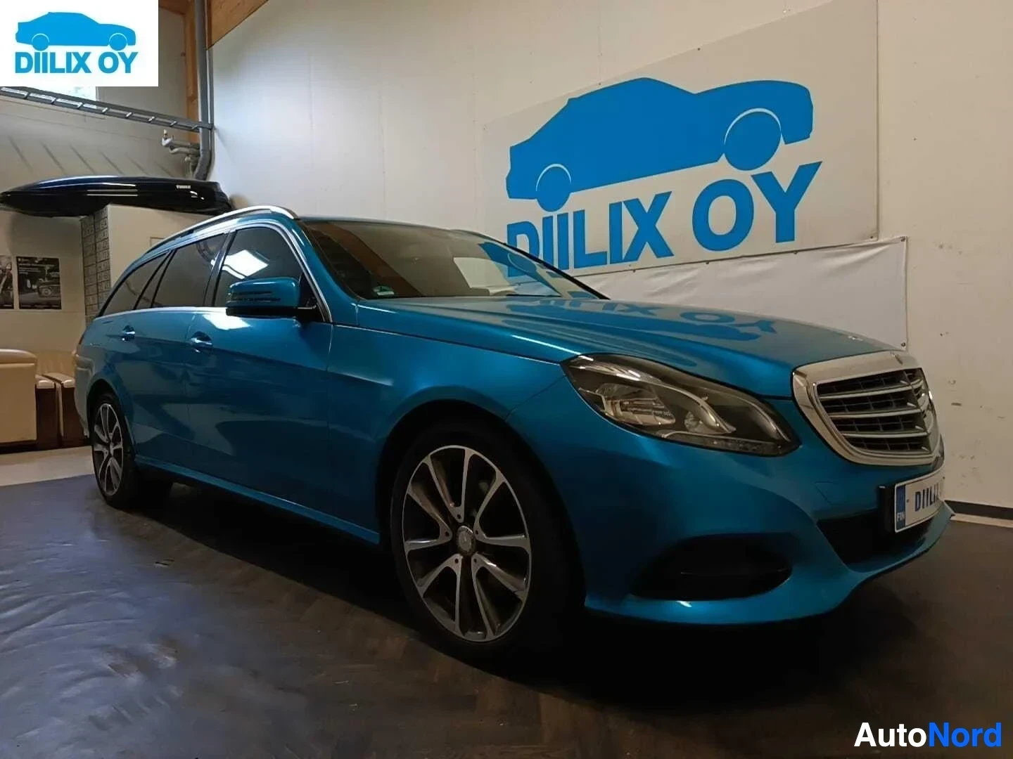 Etuviisto, nokka oikealle: Sininen Mercedes-Benz E-sarjan farmari sisällä varastossa, kuljettajaovi avoin, näkyvät mustat vanteet ja etupuoli.